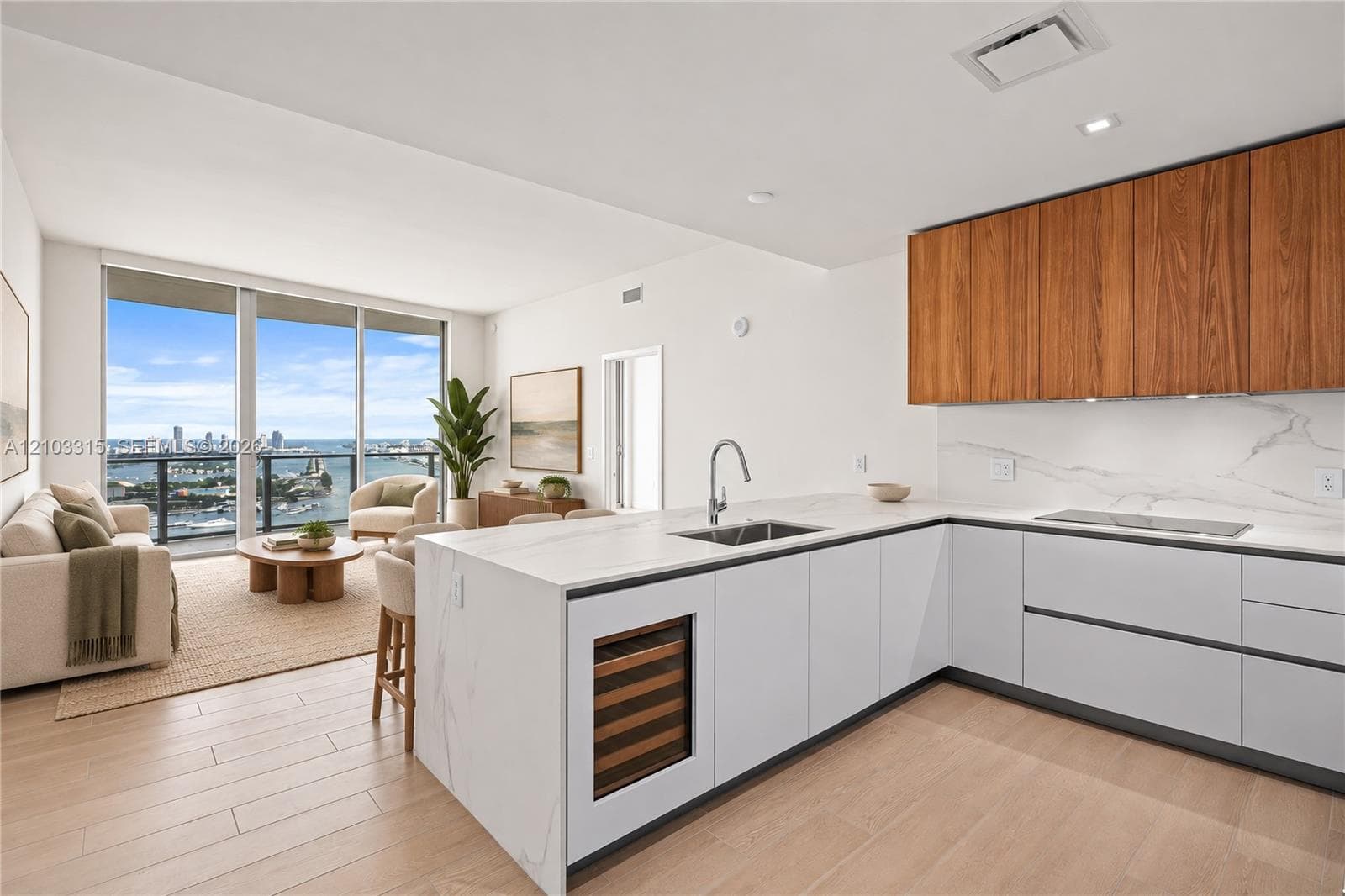Open-concept living area with wood-finish flooring, a kitchen featuring light cabinetry and a built-in wine refrigerator, a large island with a waterfall edge, and floor-to-ceiling windows