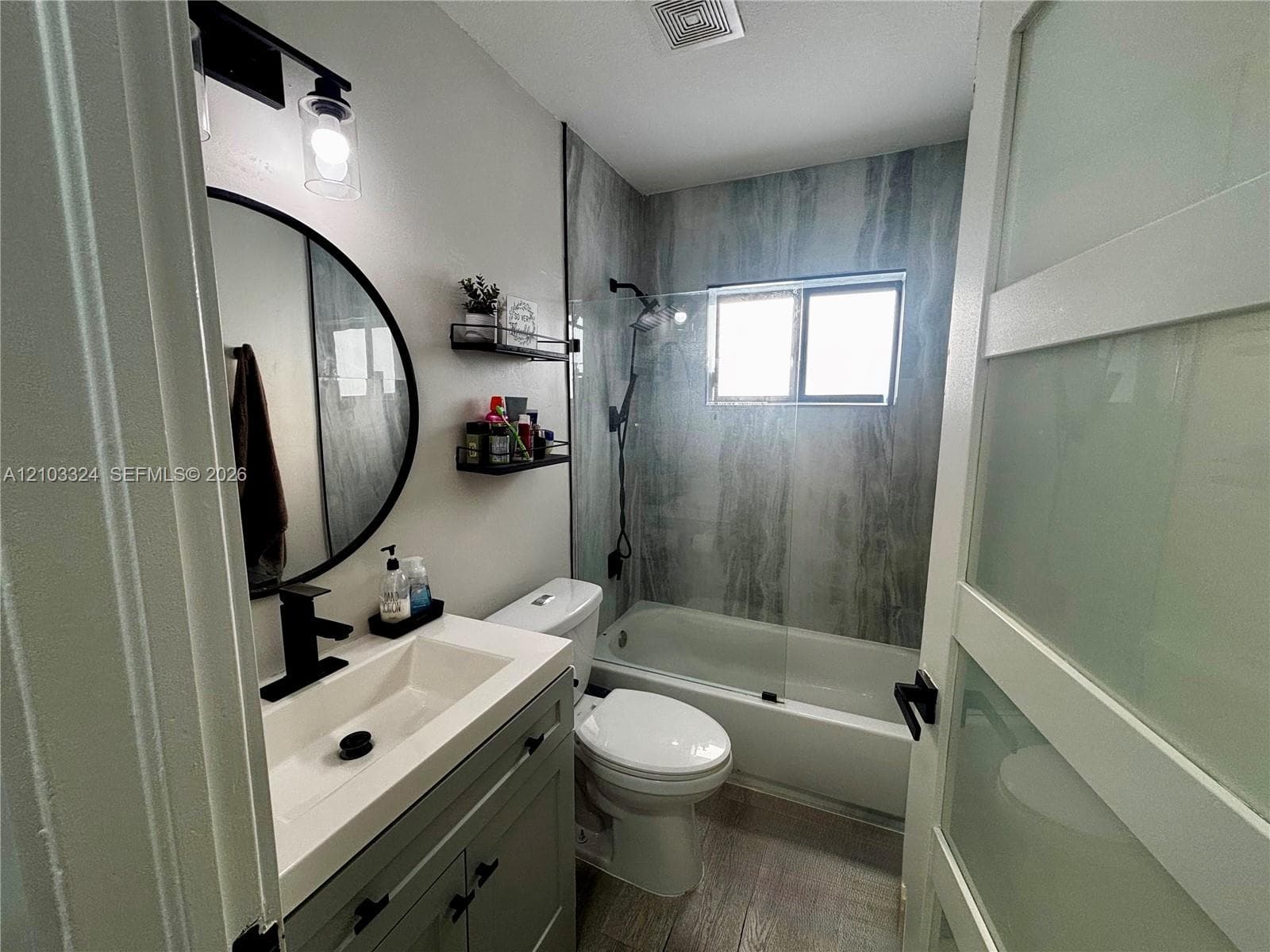 Bathroom featuring a vanity with a rectangular sink and matte black faucet, a round wall-mounted mirror, a tub-shower combination with floor-to-ceiling surround, and a glass-paneled sliding door