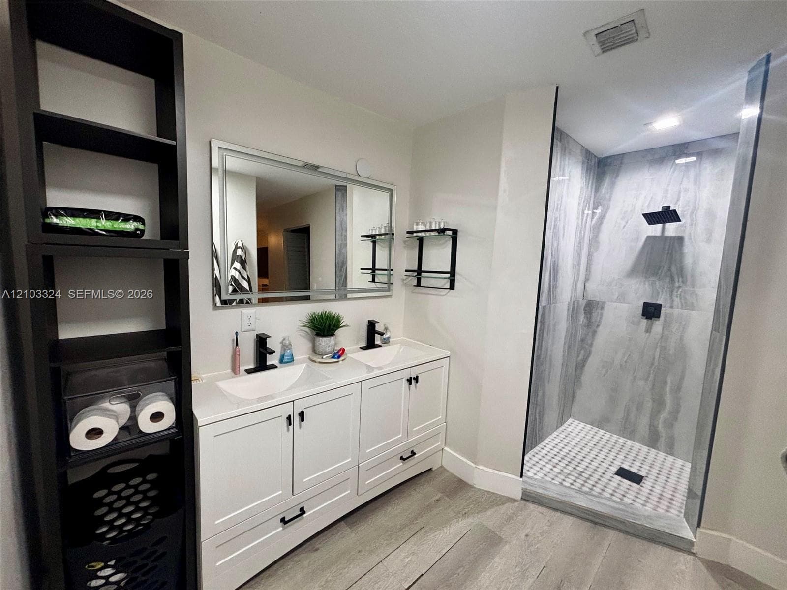 Dual-sink vanity with white cabinetry, rectangular vessel sinks, quartz countertops, and matte black fixtures, paired with a walk-in shower featuring large-format tile surround and wood-finish flooring