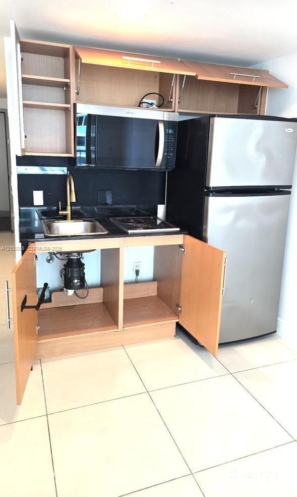 Kitchenette featuring wood-finish cabinetry, stainless steel refrigerator, over-the-range microwave, integrated cooktop, and gold-tone faucet with a drop-in sink