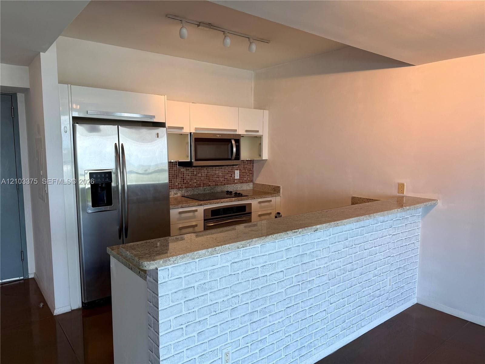 Kitchen featuring white flat-panel cabinetry, stainless steel appliances, stone-surface breakfast bar with a white brick base, and mosaic tile backsplash