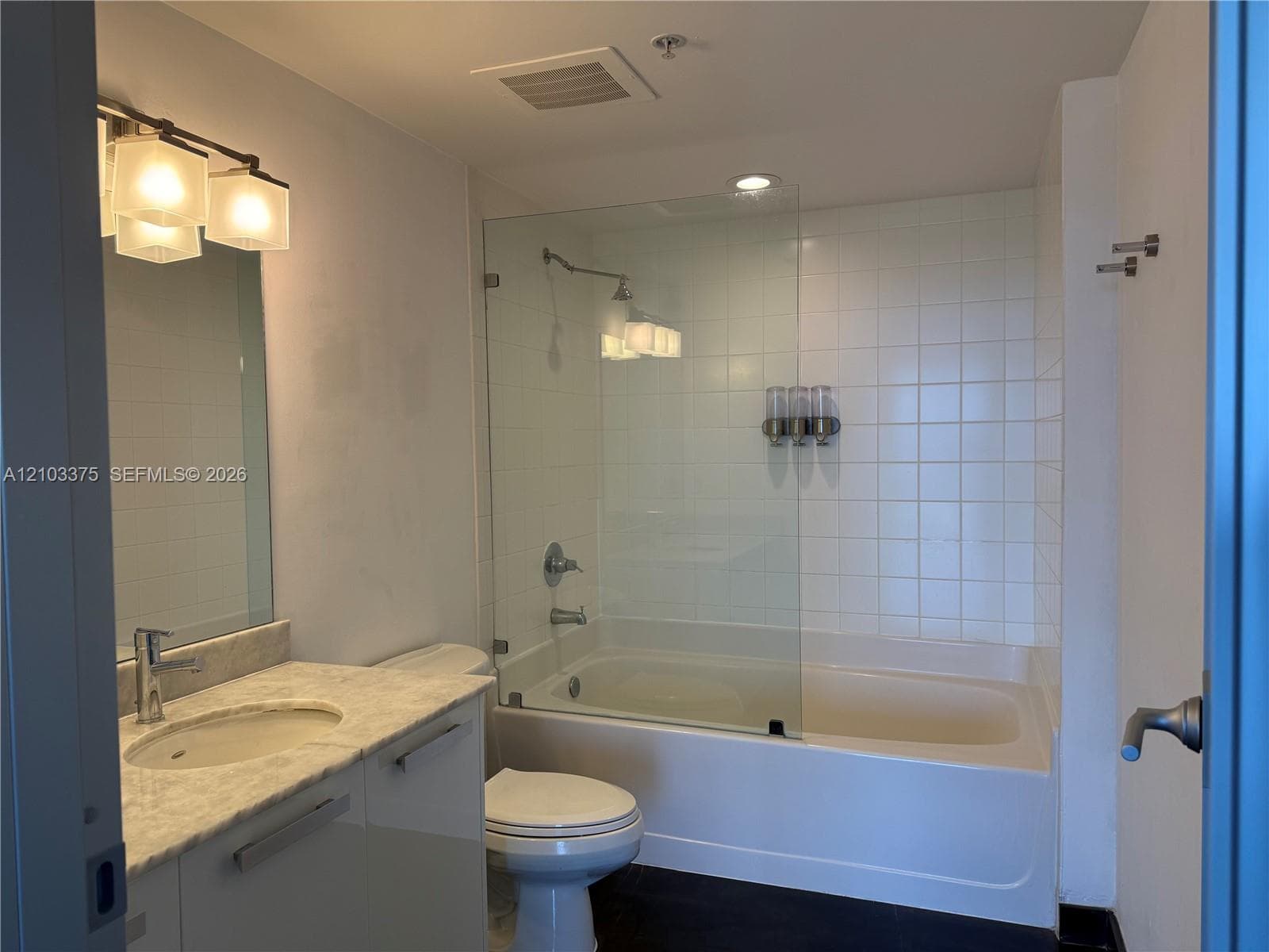Bathroom featuring a stone-top vanity with an integrated sink, frameless glass shower panel, white tile tub surround, and three-light wall-mounted fixture