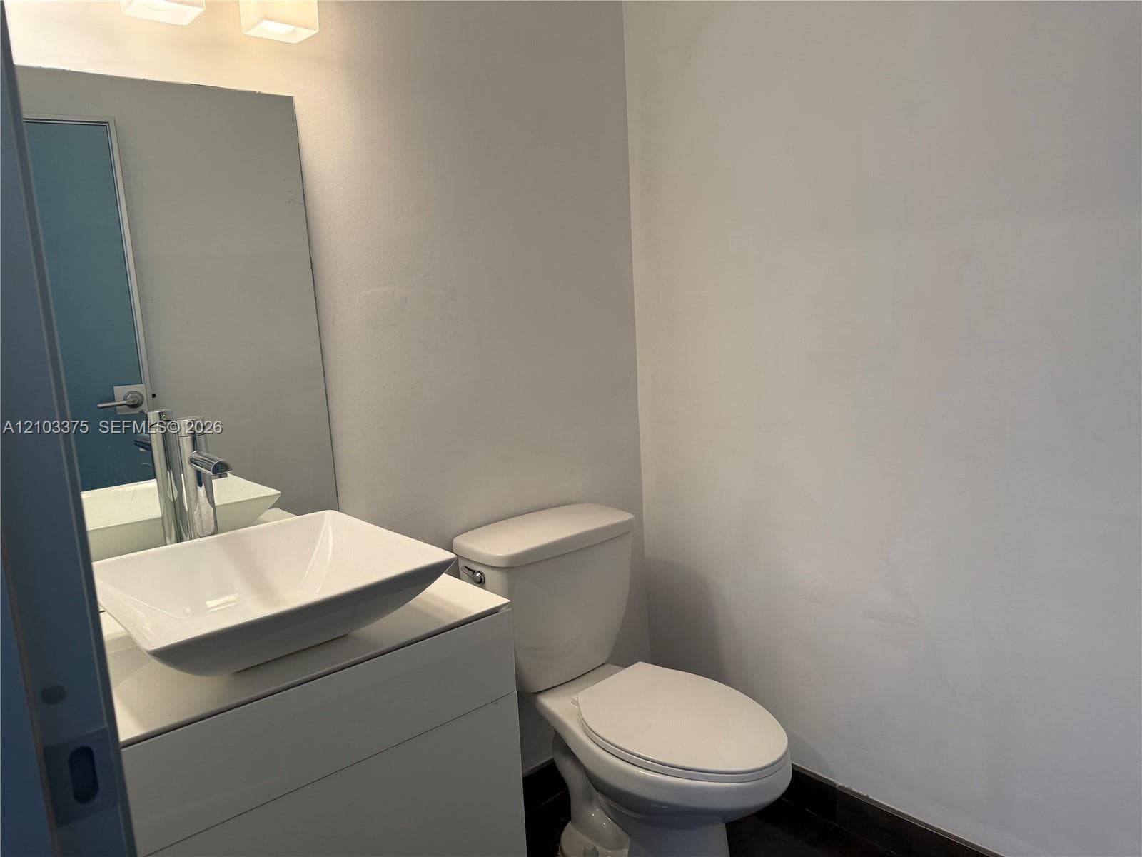 White vanity with a rectangular vessel sink and a single-handle chrome faucet, paired with a frameless wall mirror, overhead lighting fixture, and a standard toilet