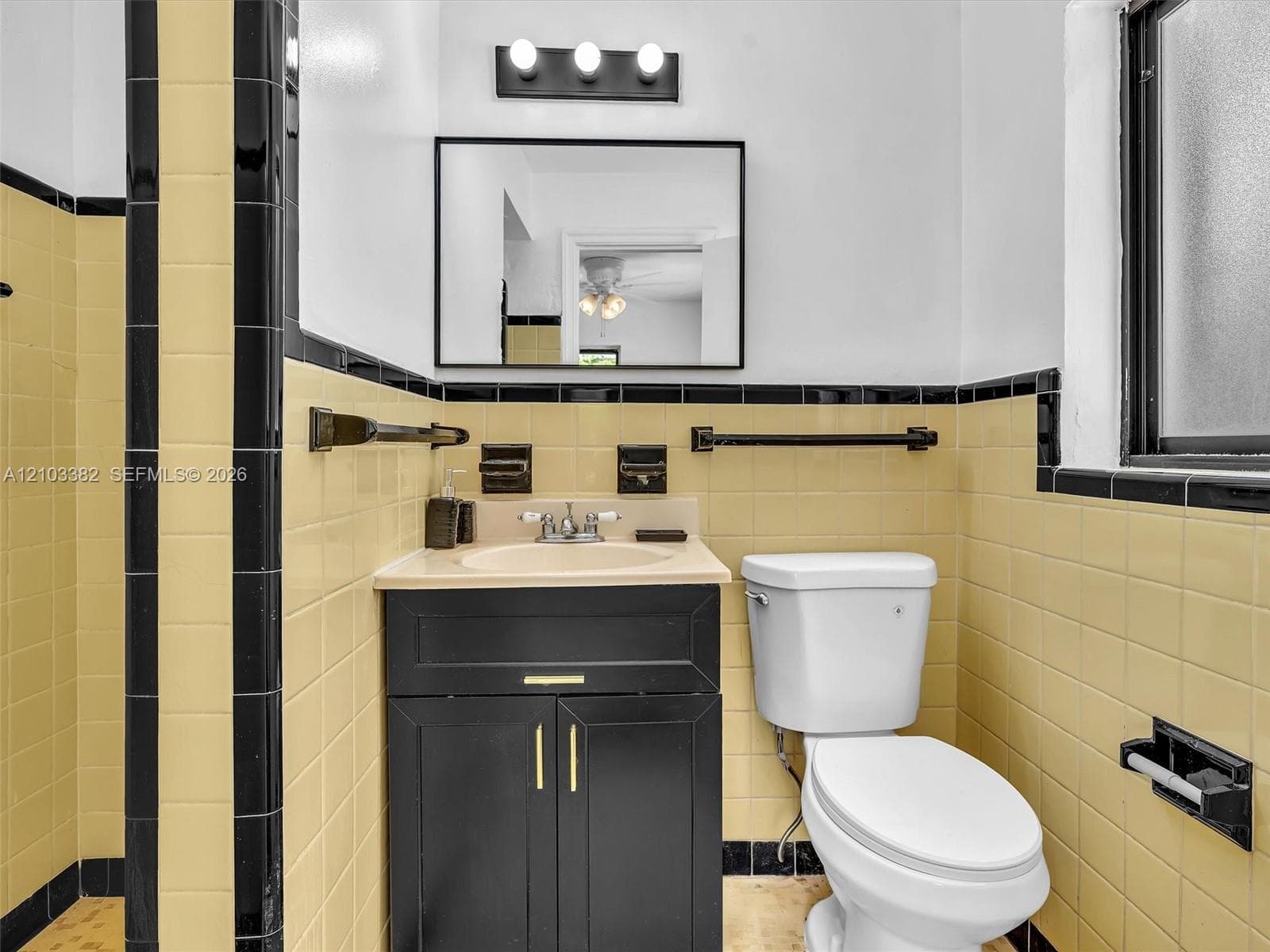 Black vanity cabinet with brass-tone hardware, rectangular wall-mounted mirror, three-light fixture, and ceramic tile wall accents featuring contrasting border detailing. House for rent in Coral Gables. House for rent listed by Realtor.