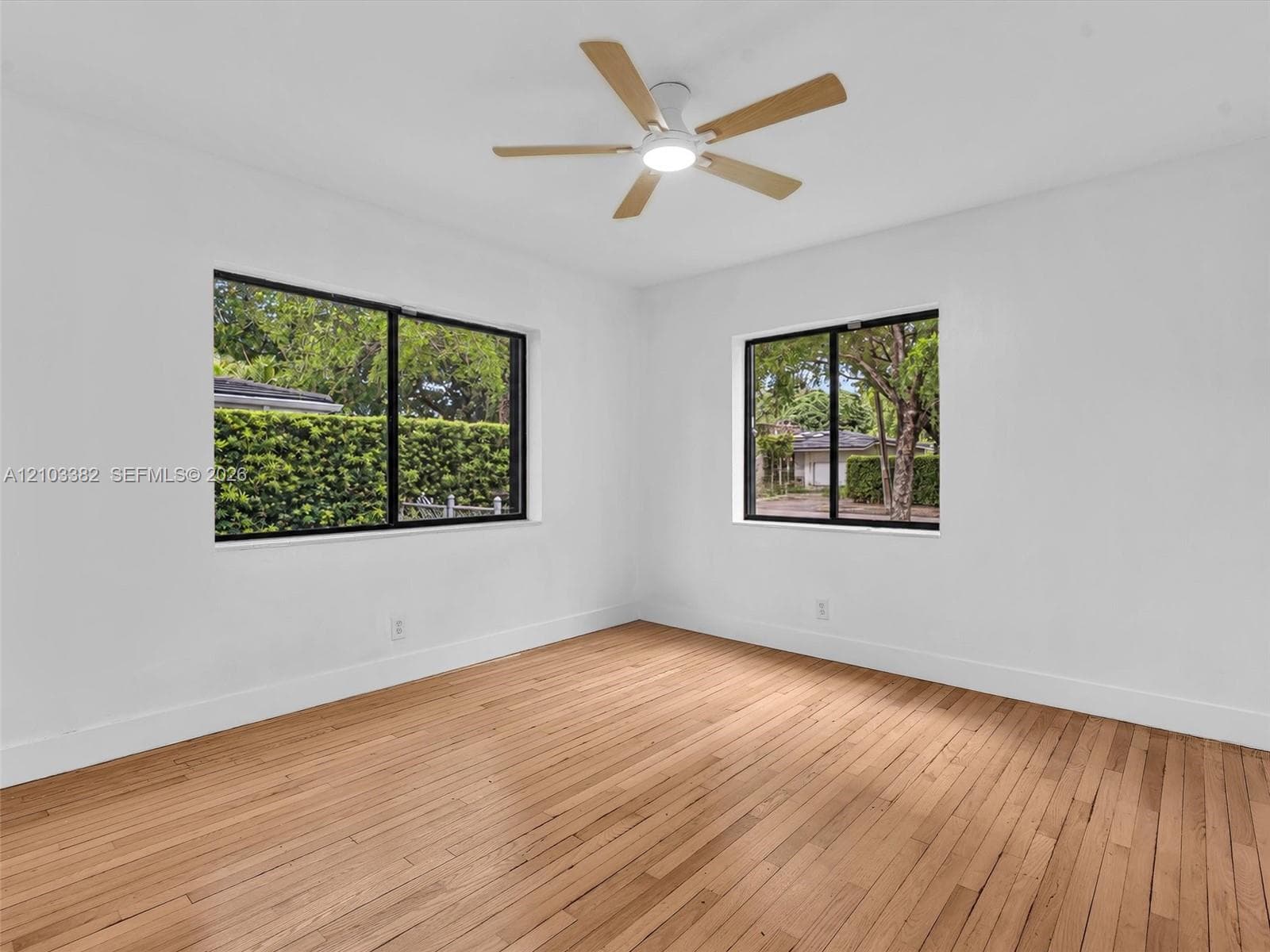 Large front/east bedroom with sunny windows. Featuring wood-finish flooring, two windows with black frames, and a multi-blade ceiling fan with integrated lighting. House for rent in Coral Gables. House for rent listed by Realtor.