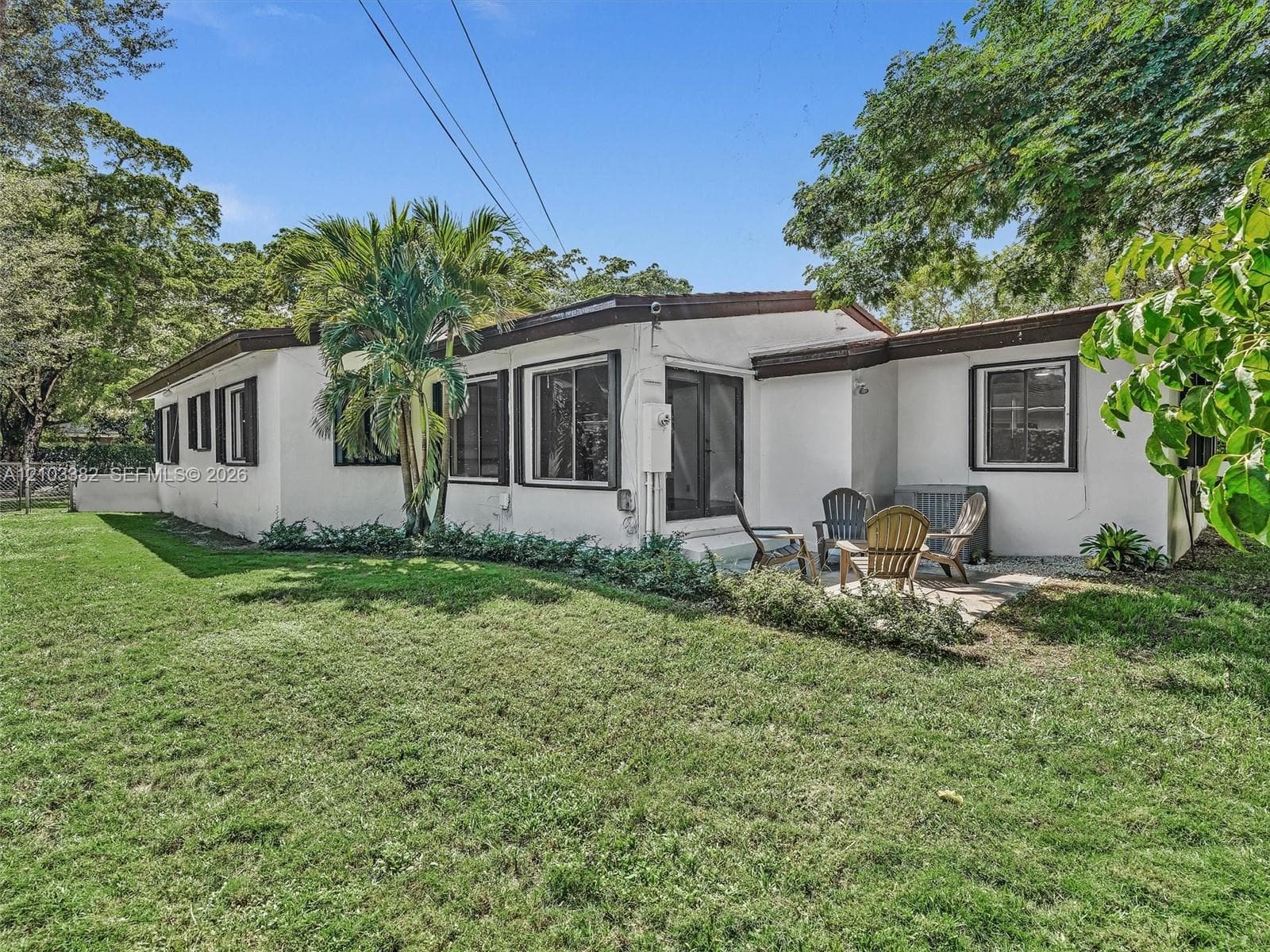 Exterior featuring white stucco siding, dark-trimmed windows, a concrete patio, and a fenced grass lawn with mature trees and palm foliage. House for rent in Coral Gables. House for rent listed by Realtor.