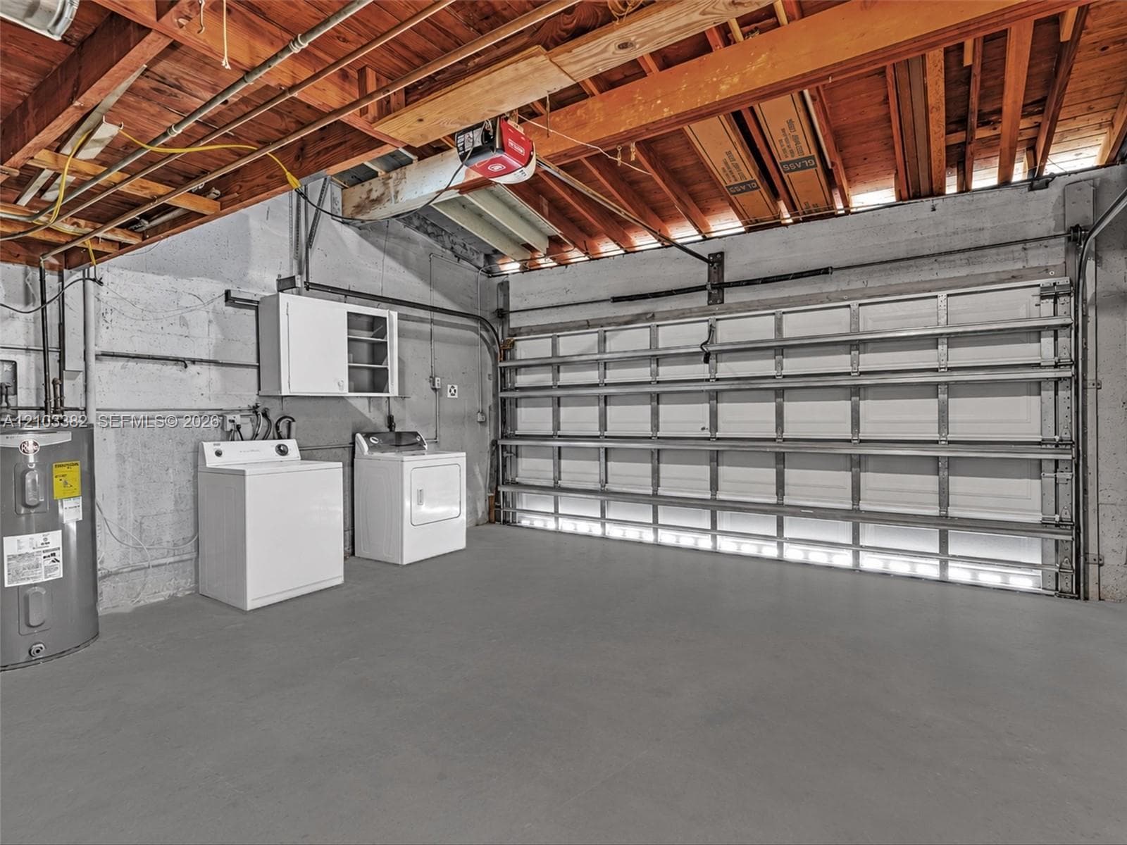 Huge 2 car, attached garage featuring a white sectional roll-up door, exposed wood joist ceiling, concrete walls, and concrete flooring. House for rent in Coral Gables. House for rent listed by Realtor.