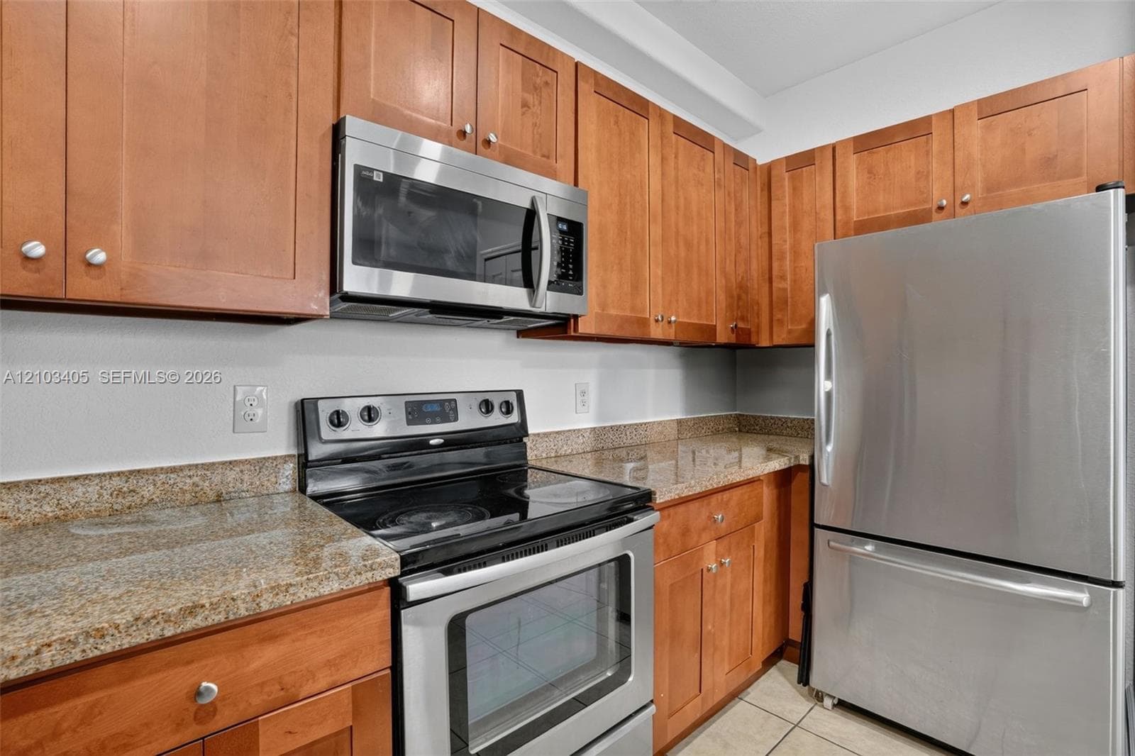 Kitchen featuring wood cabinetry, stone countertops, stainless steel dual-basin undermount sink with a gooseneck faucet, stainless steel refrigerator, and tile flooring