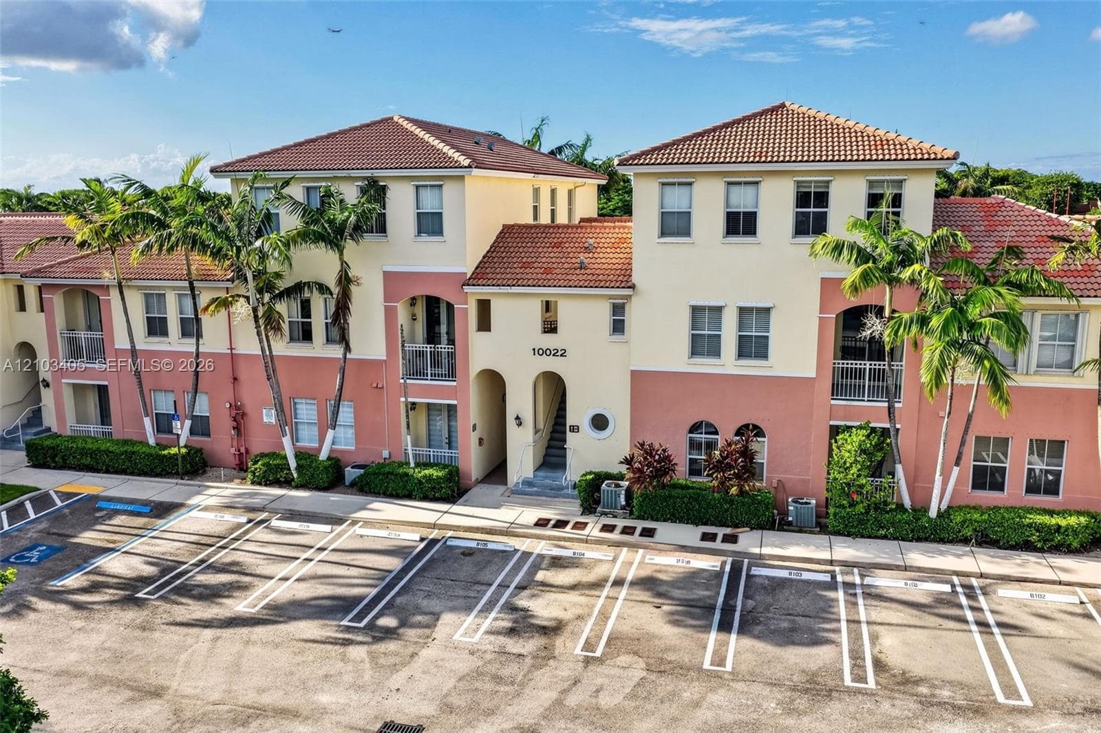 Aerial perspective of multi-story residential buildings featuring tile roofs, lakeside positioning, paved pathways, and mature palm tree landscaping