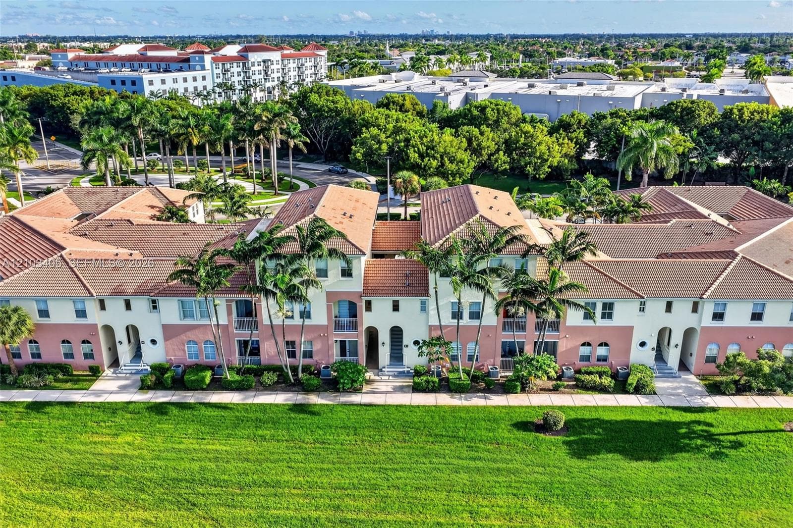 Aerial perspective of a multi-unit residential complex featuring tile roofing, stucco exterior, paved parking areas, surrounding greenbelts, and a water feature