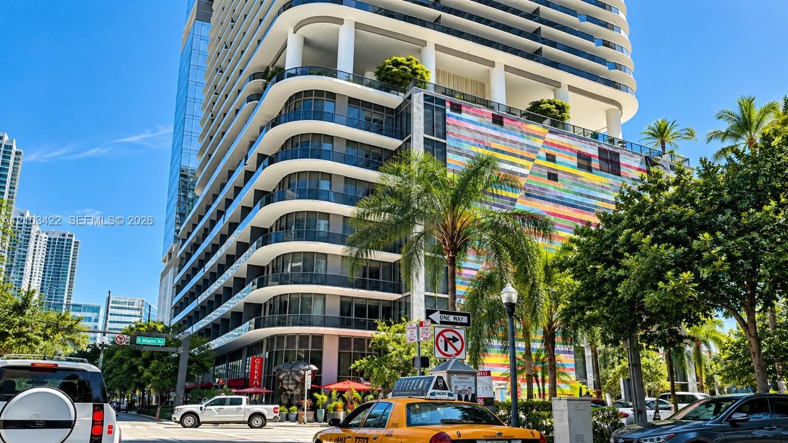 Modern high-rise building with curved balconies, a vibrant multi-color facade, street-level retail spaces, and surrounding mature palm trees
