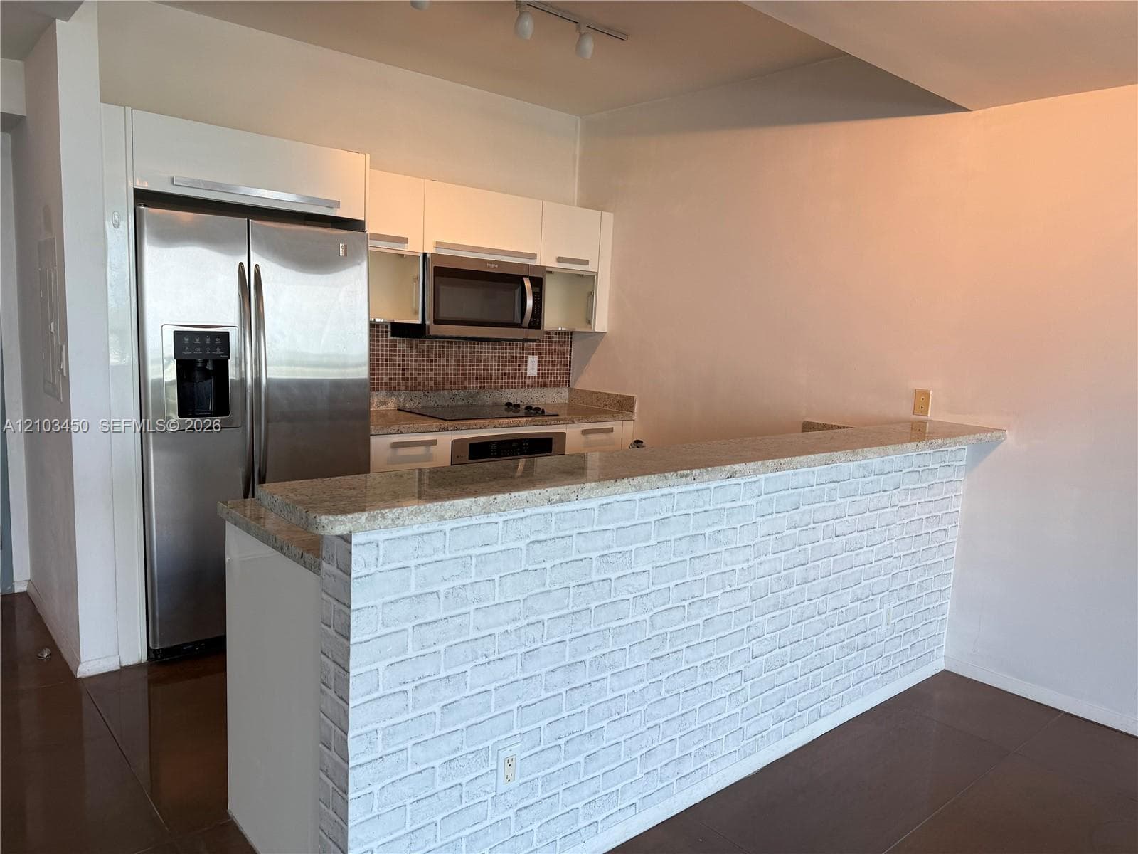 Kitchen featuring white cabinetry, stainless steel refrigerator, built-in microwave, stone-surface countertop, and a white brick-finish peninsula
