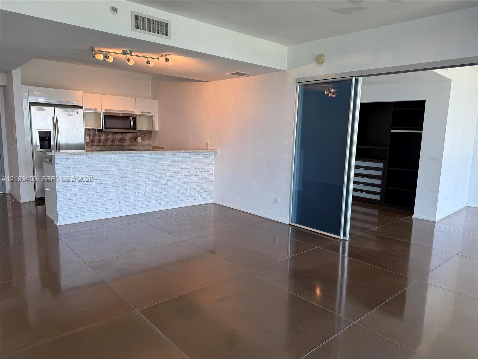 Kitchen featuring white cabinetry, stainless steel appliances, a breakfast bar with textured stone-style cladding, large-format tile flooring, and a sliding glass partition with integrated closet shelving
