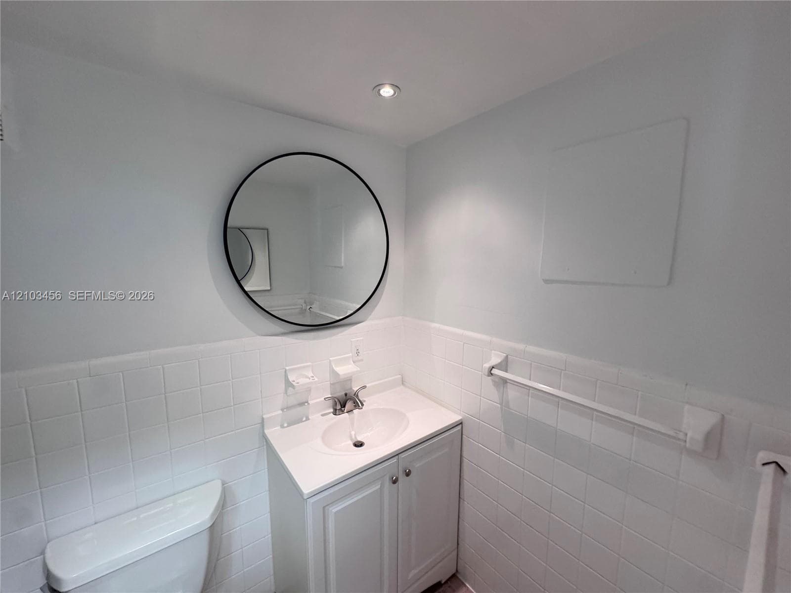 White vanity with a molded countertop and integrated basin, circular wall-mounted mirror with a black frame, recessed lighting, and white ceramic wall tiling