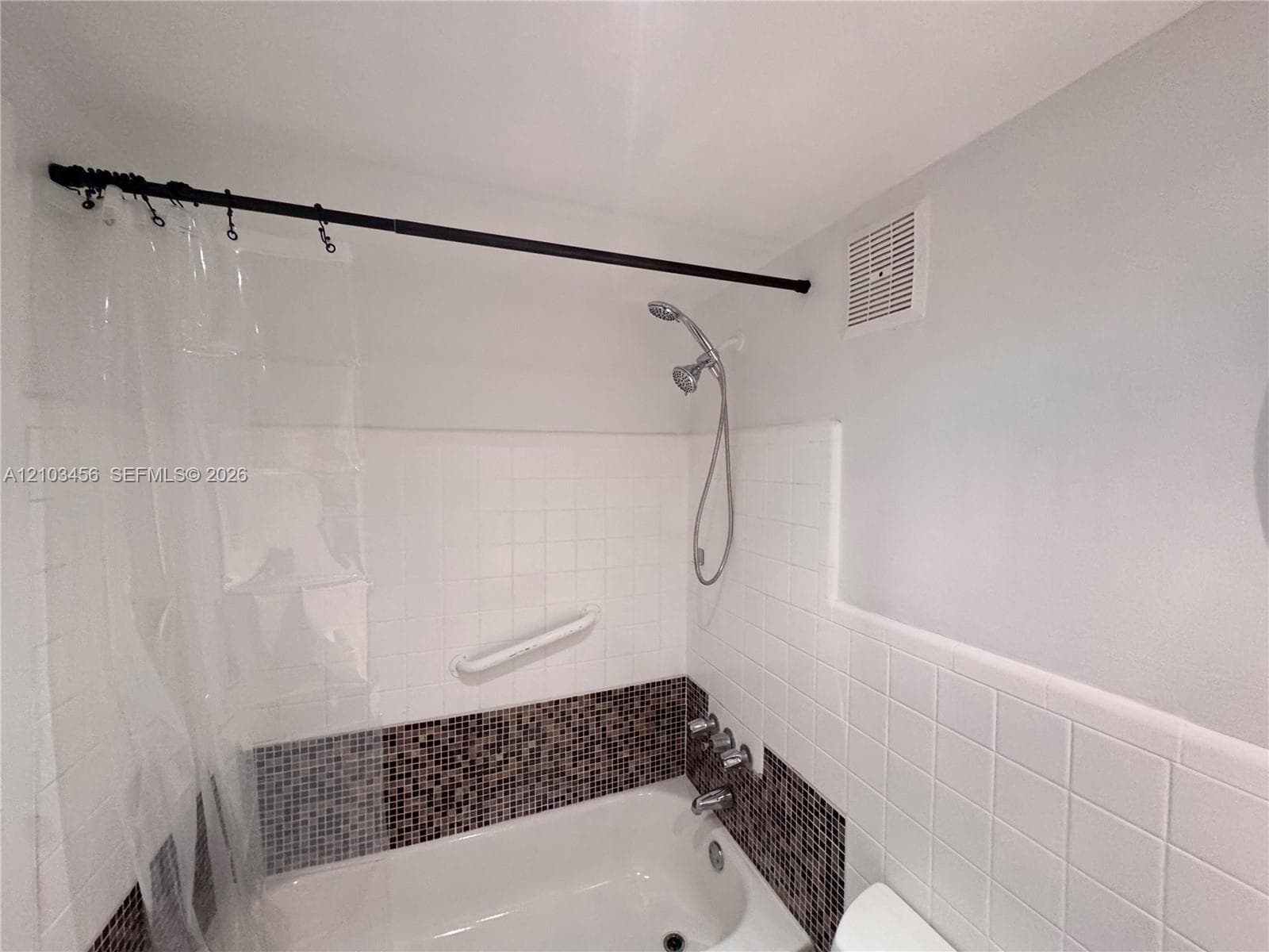 Bathtub featuring white square wall tile, decorative mosaic accent tile, handheld showerhead, and a wall-mounted grab bar