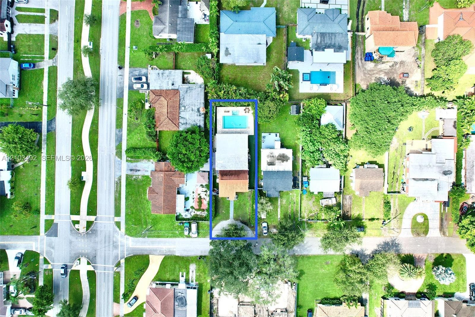 Aerial perspective featuring a rectangular swimming pool, concrete paved patio area, front driveway, and mature landscaping on a fenced lot