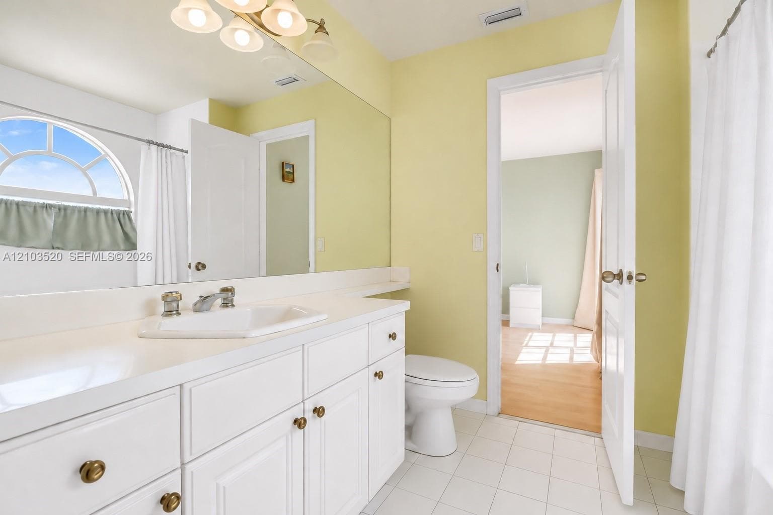 Bathroom featuring a white vanity with integrated sink, expansive wall-mounted mirror, polished chrome faucet, arched window, and ceramic tile flooring