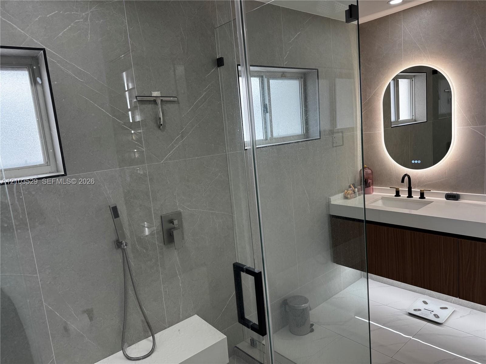 Walk-in shower with floor-to-ceiling large-format tile, frameless glass enclosure, wall-mounted shower fixture, floating wood-finish vanity, and integrated backlit oval mirror