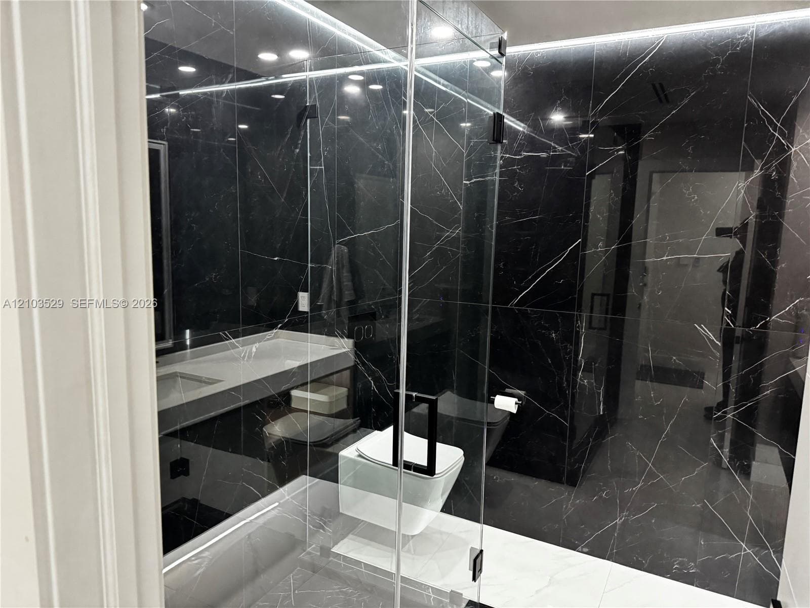 Walk-in glass-enclosed shower featuring dark, veined tile walls, wall-mounted white toilet, integrated lighting, and floating vanity with a white composite countertop