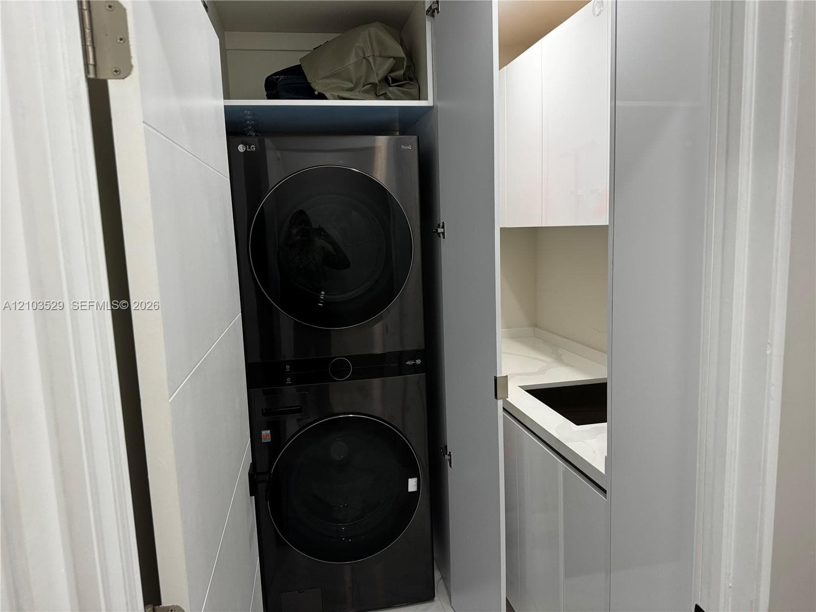 Laundry alcove featuring a stacked appliance unit, high-gloss white cabinetry, solid-surface countertops, and an integrated basin