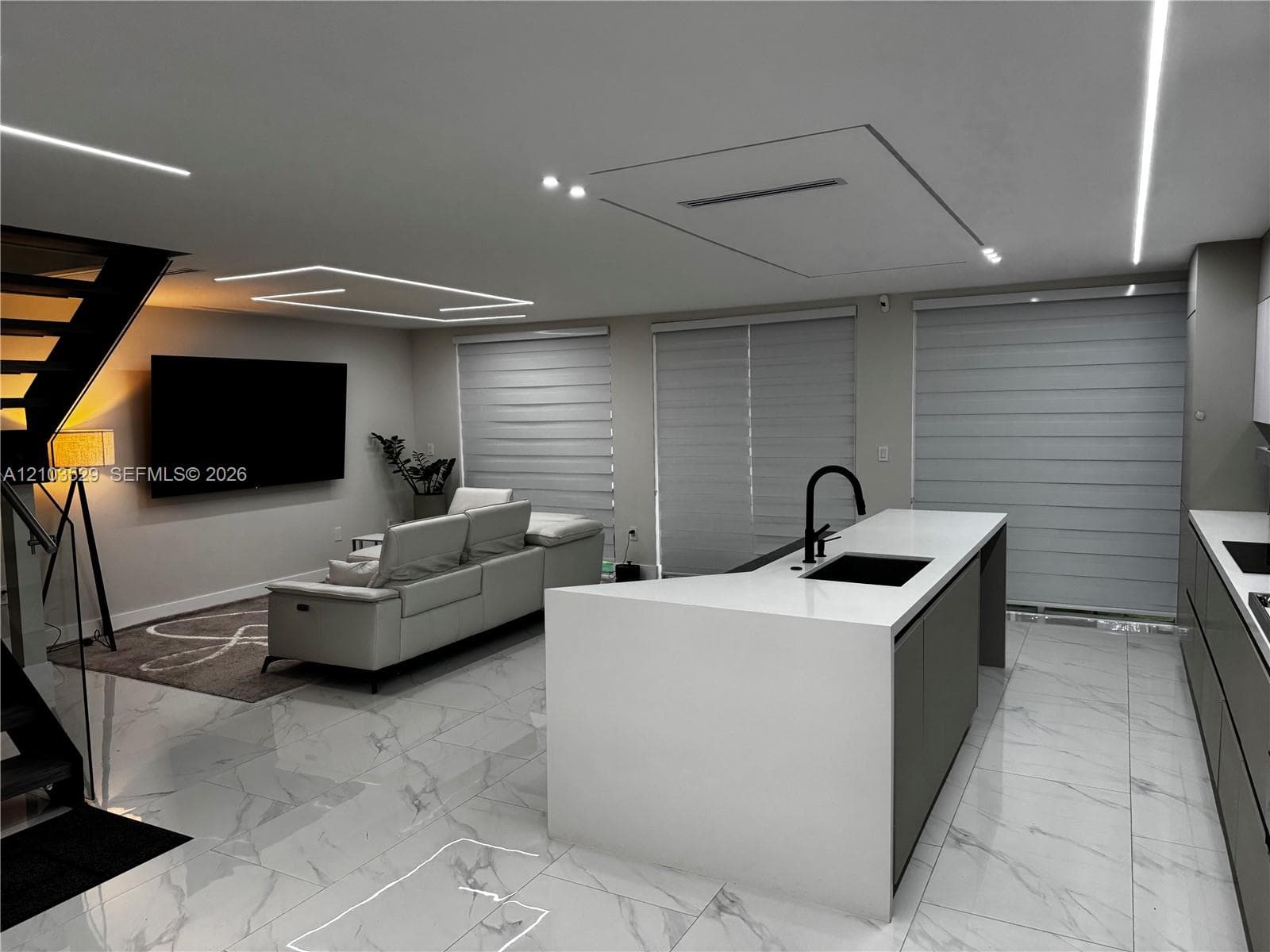Large-format tile flooring, kitchen island with a neutral countertop and integrated sink, matte black plumbing fixtures, integrated linear ceiling lighting, and metal open-riser staircase