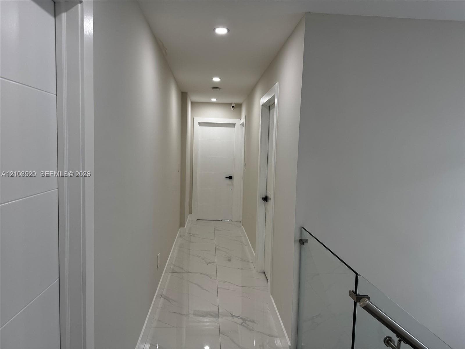 Hallway featuring large-format tile flooring with a neutral veined pattern, recessed ceiling lighting, glass panel railing with metallic hardware, and minimalist interior doors with matte black levers