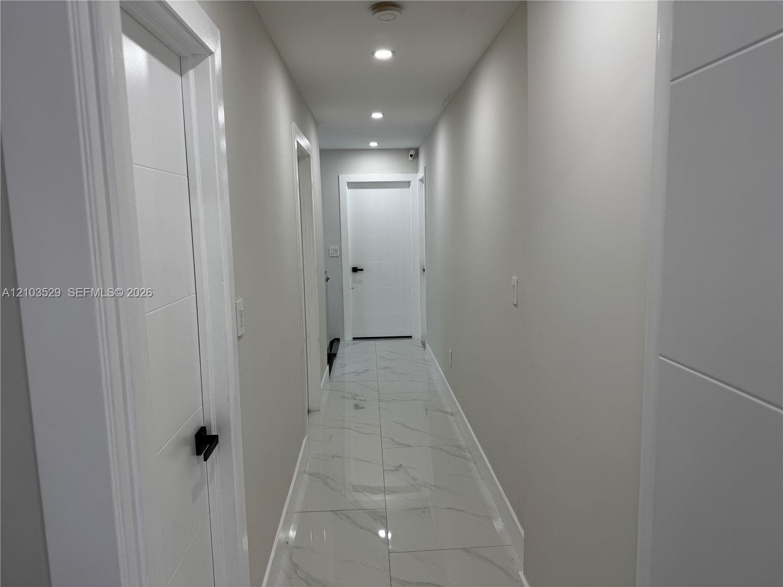 Hallway featuring large-format tile flooring with a neutral veined pattern, recessed ceiling lighting, and solid-core paneled doors equipped with matte black hardware
