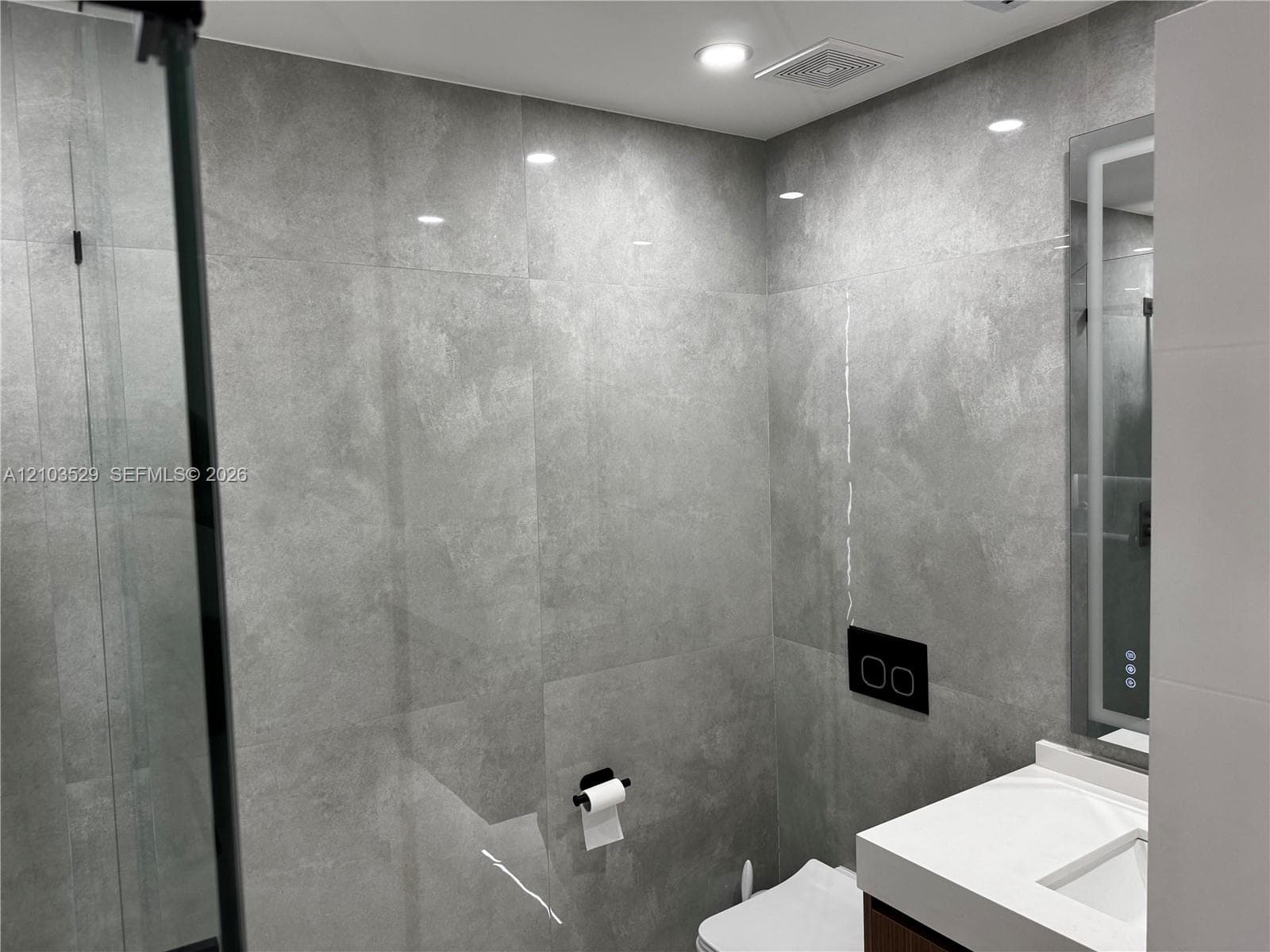 Full-height grey large-format wall tiling, wall-mounted dual-flush toilet, vanity with integrated sink and countertop, and LED-backlit mirror