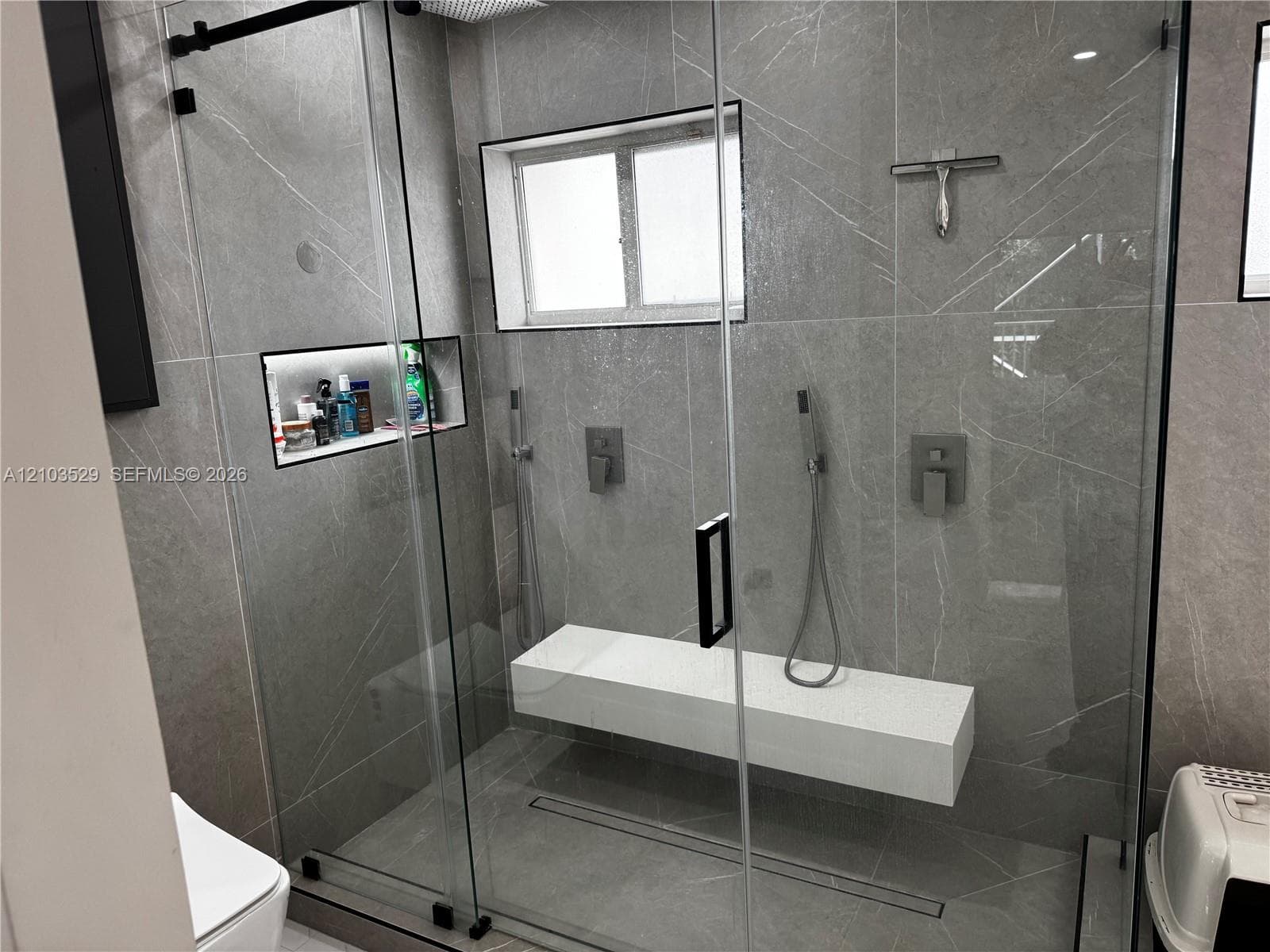 Walk-in shower featuring large-format tile walls, a sliding glass door, a floating stone bench, recessed wall shelving, and dual wall-mounted shower fixtures