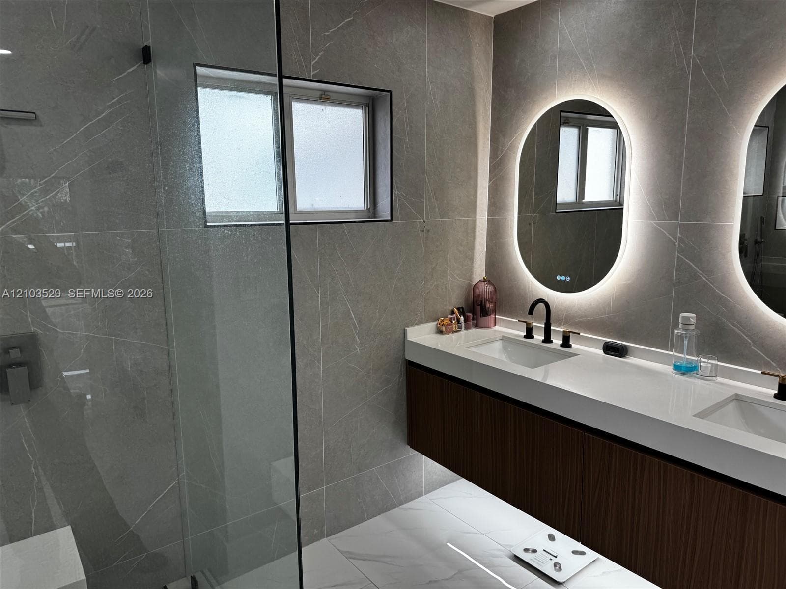 Floating double vanity with dual undermount sinks, quartz countertop, backlit oval mirrors, floor-to-ceiling large-format tile wall cladding, and a frameless glass-enclosed shower