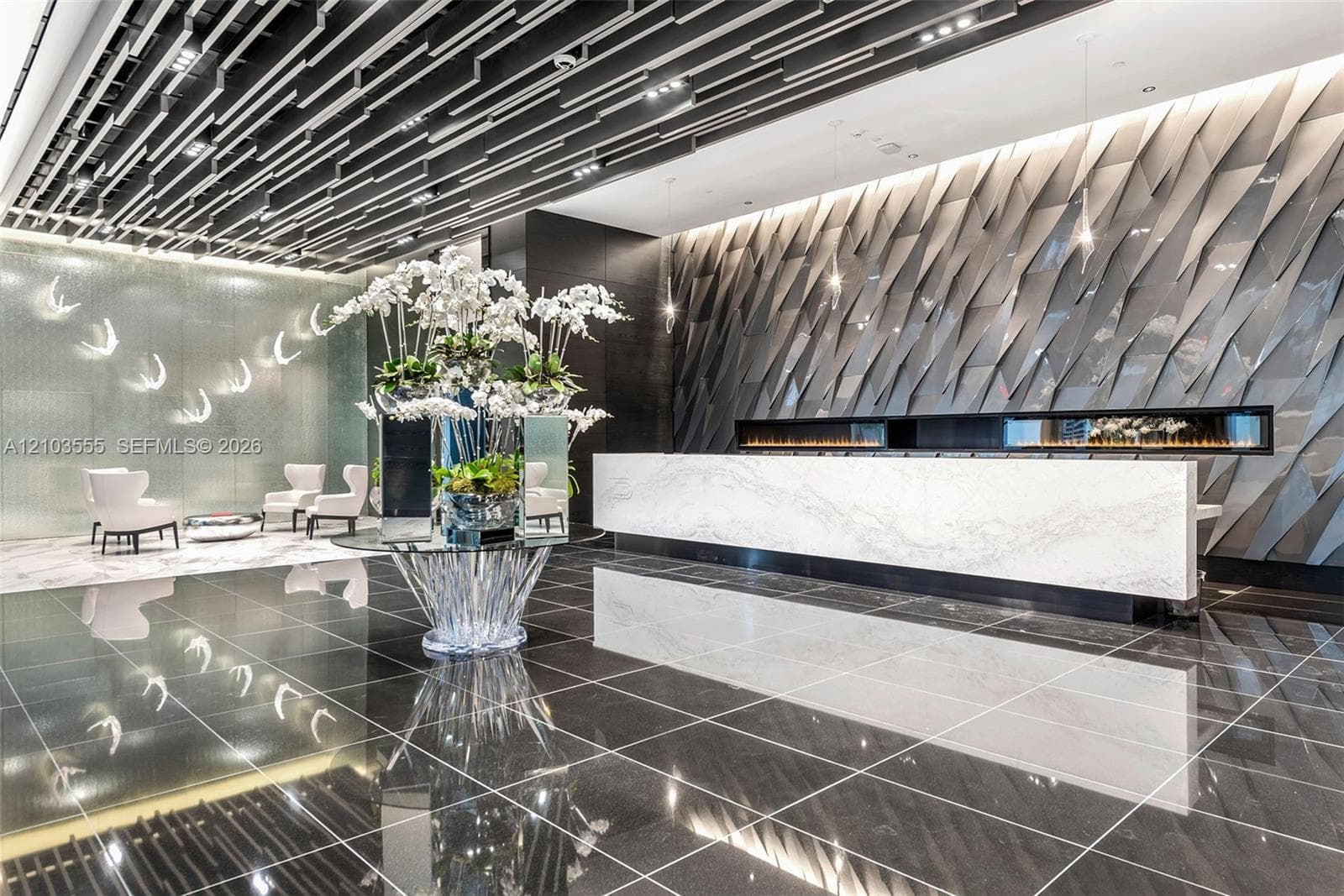 Reflective dark tile flooring, a marble-finish reception desk, a linear fireplace, an illuminated decorative wall panel, and a striking ceiling design