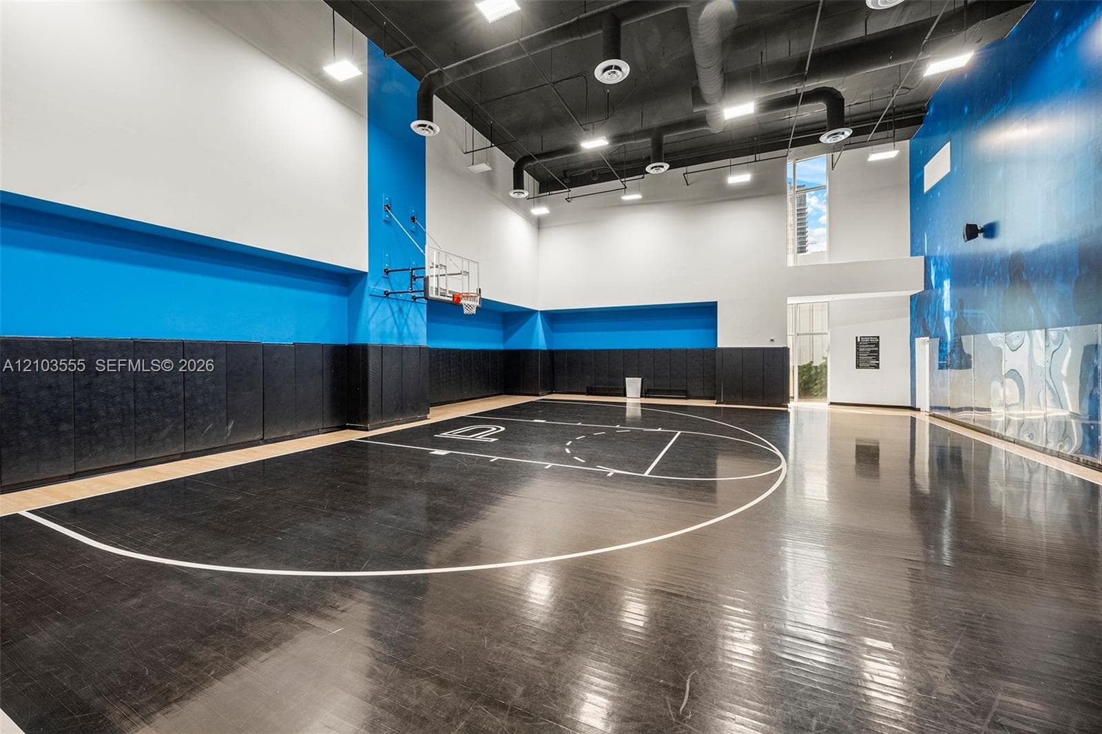 Indoor sports court featuring a dark wood-finish floor with court markings, padded wall protection, high ceilings, and recessed lighting