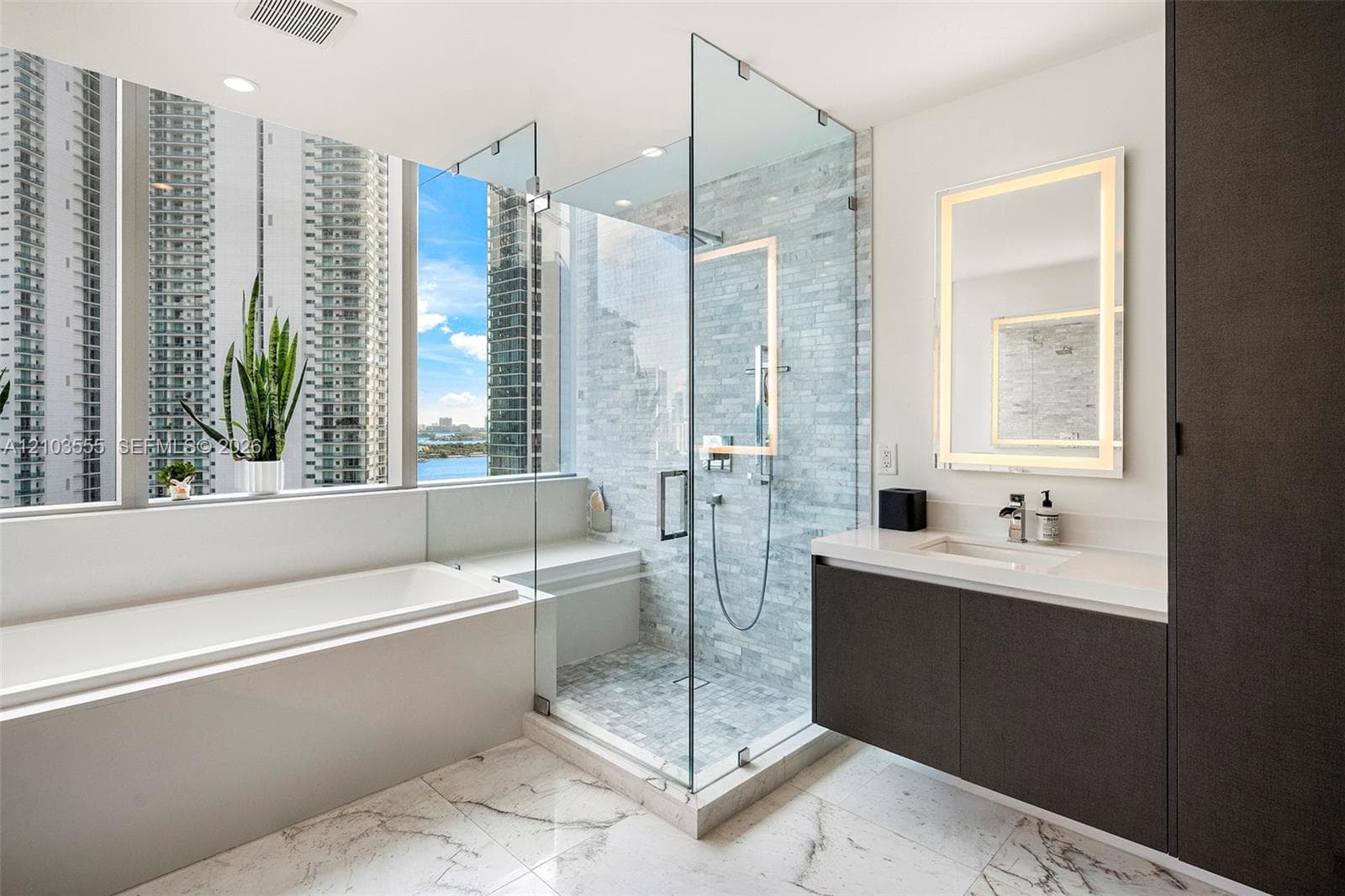 Floor-to-ceiling window, standalone soaking tub, glass-enclosed shower, floating vanity with integrated sink, and illuminated mirror