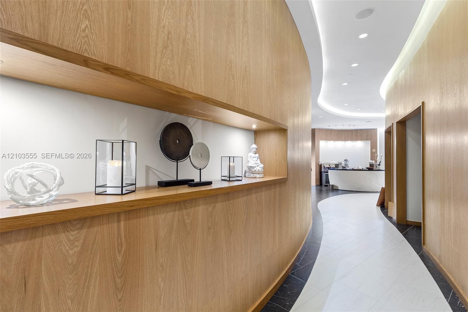 Wood-finish wall paneling with integrated display niche, recessed ceiling lighting, and a curved hallway design with contrasting floor tiling