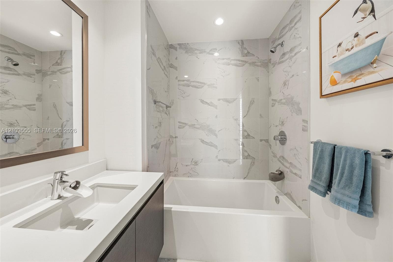 Integrated bathtub and shower with stone-look tile surround, single vanity with solid surface countertop, undermount sink, and wall-mounted mirror