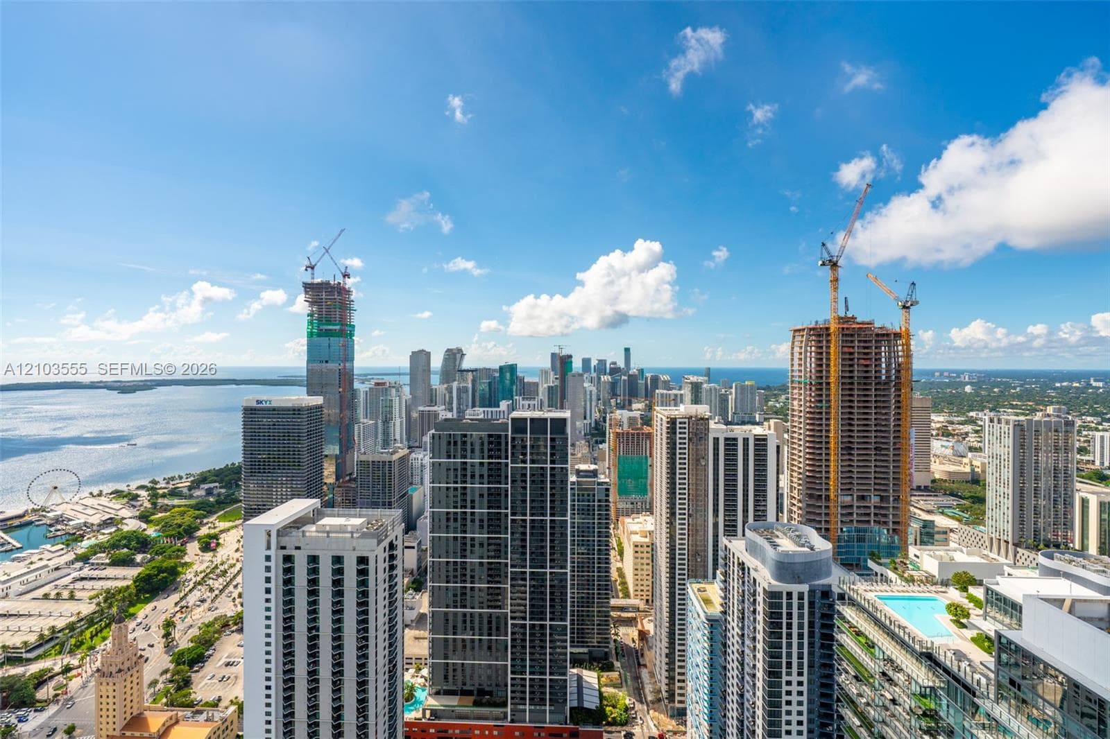 Expansive cityscape with a prominent body of water, multiple high-rise buildings, and a clear sky with scattered clouds