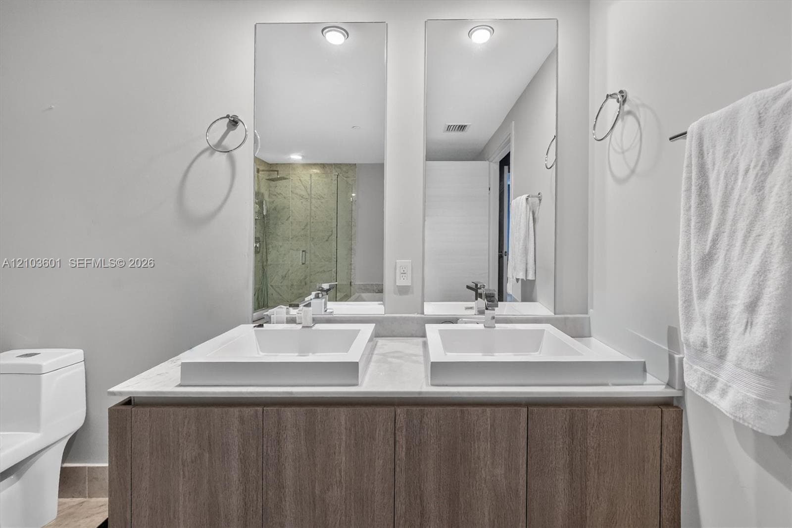 Dual vanity featuring wood-finish cabinetry, stone-surface countertop, rectangular vessel sinks, and wall-mounted vanity mirrors with integrated lighting