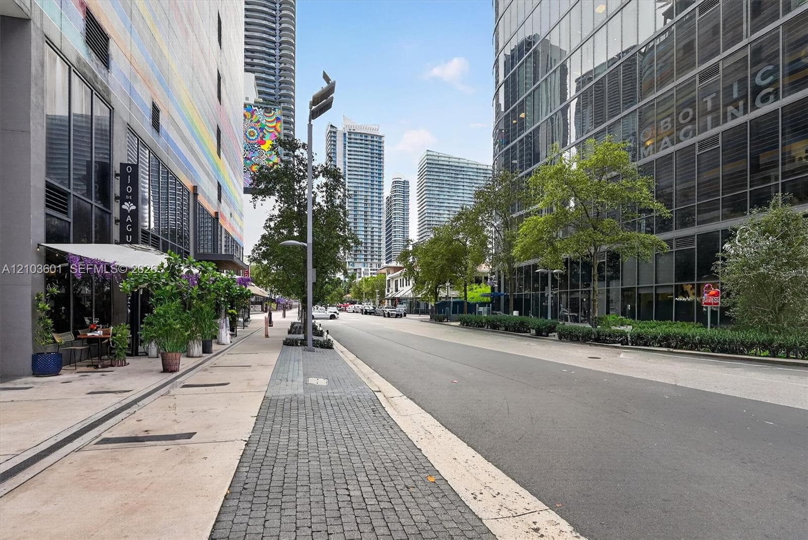 Urban streetscape featuring paved sidewalks, cobblestone accents, mature trees, and high-rise glass architecture