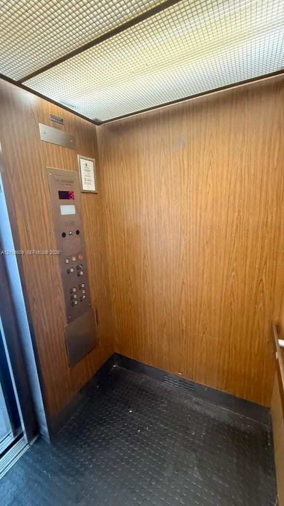 Elevator interior featuring wood-finish paneling, grid-patterned ceiling lighting, metallic control panel, and textured flooring