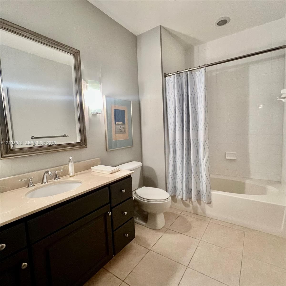 Bathroom featuring dark-finish cabinetry, light-toned stone-style countertop, integrated sink with polished chrome hardware, framed vanity mirror, and tile flooring