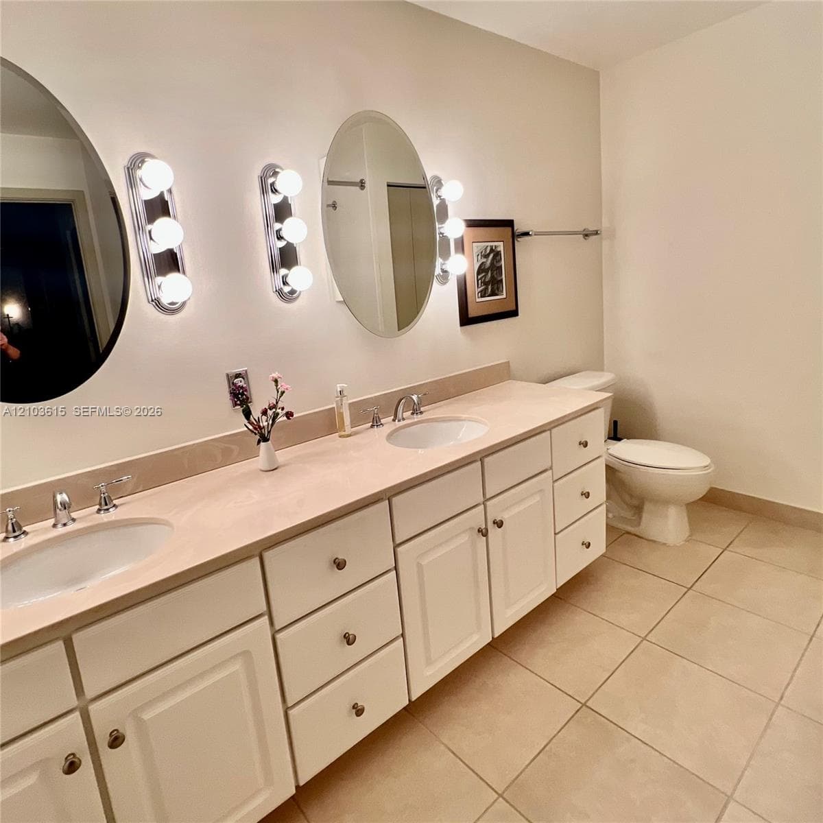 Dual-sink vanity featuring white cabinetry, a solid surface countertop, chrome-finish widespread faucets, oval wall mirrors, and three-bulb vanity lighting