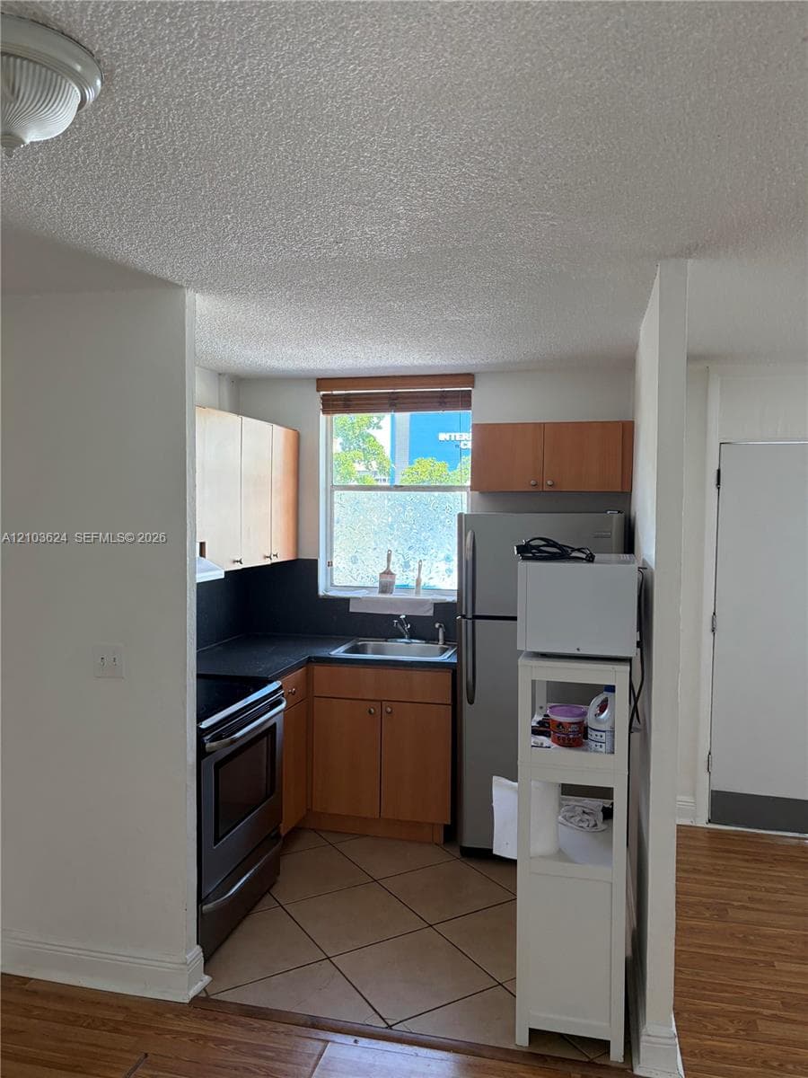 Kitchen featuring wood-finish cabinetry, dark solid-surface countertops, tile flooring, stainless steel appliance suite, and a window above the sink