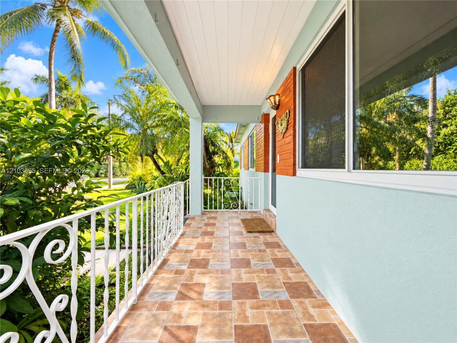 Covered porch featuring multi-tonal ceramic tile flooring, decorative wrought-iron railing, beadboard ceiling, and brick exterior accents