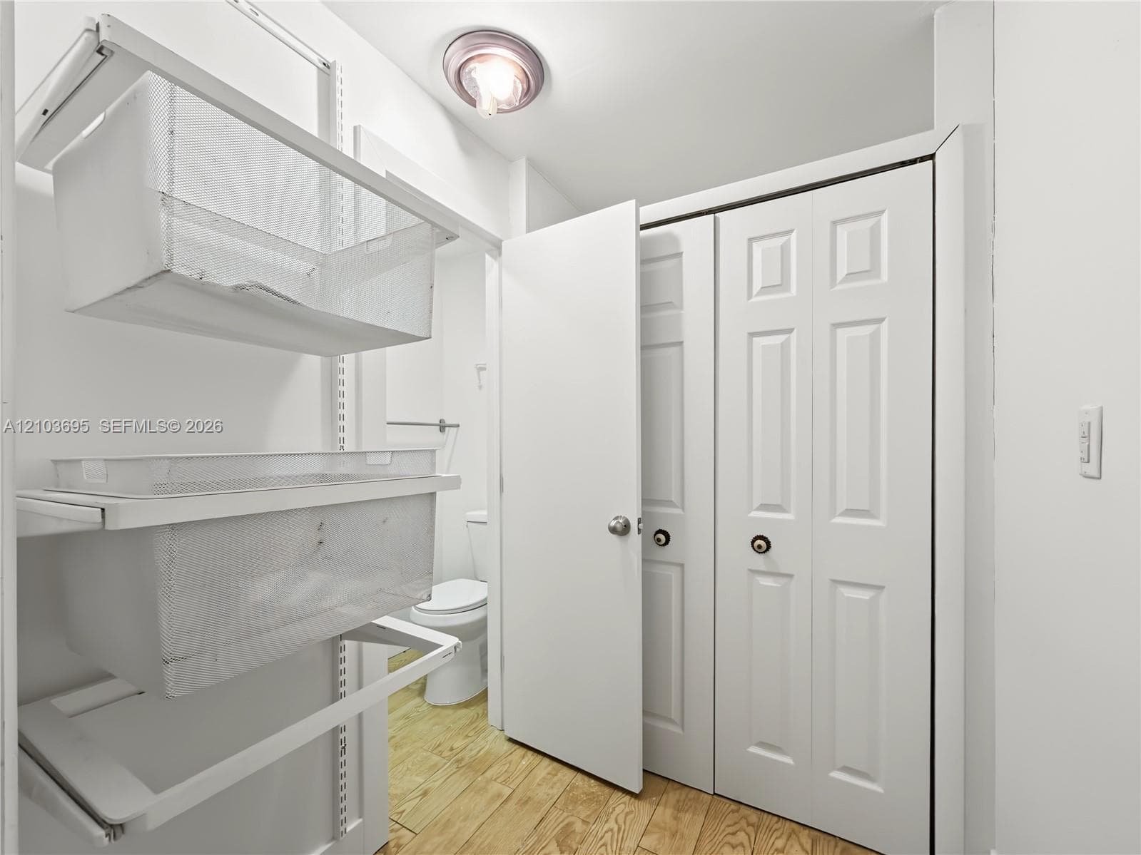 Wood-finish flooring, white paneled closet doors, and wall-mounted metal wire shelving system with flush-mount ceiling lighting