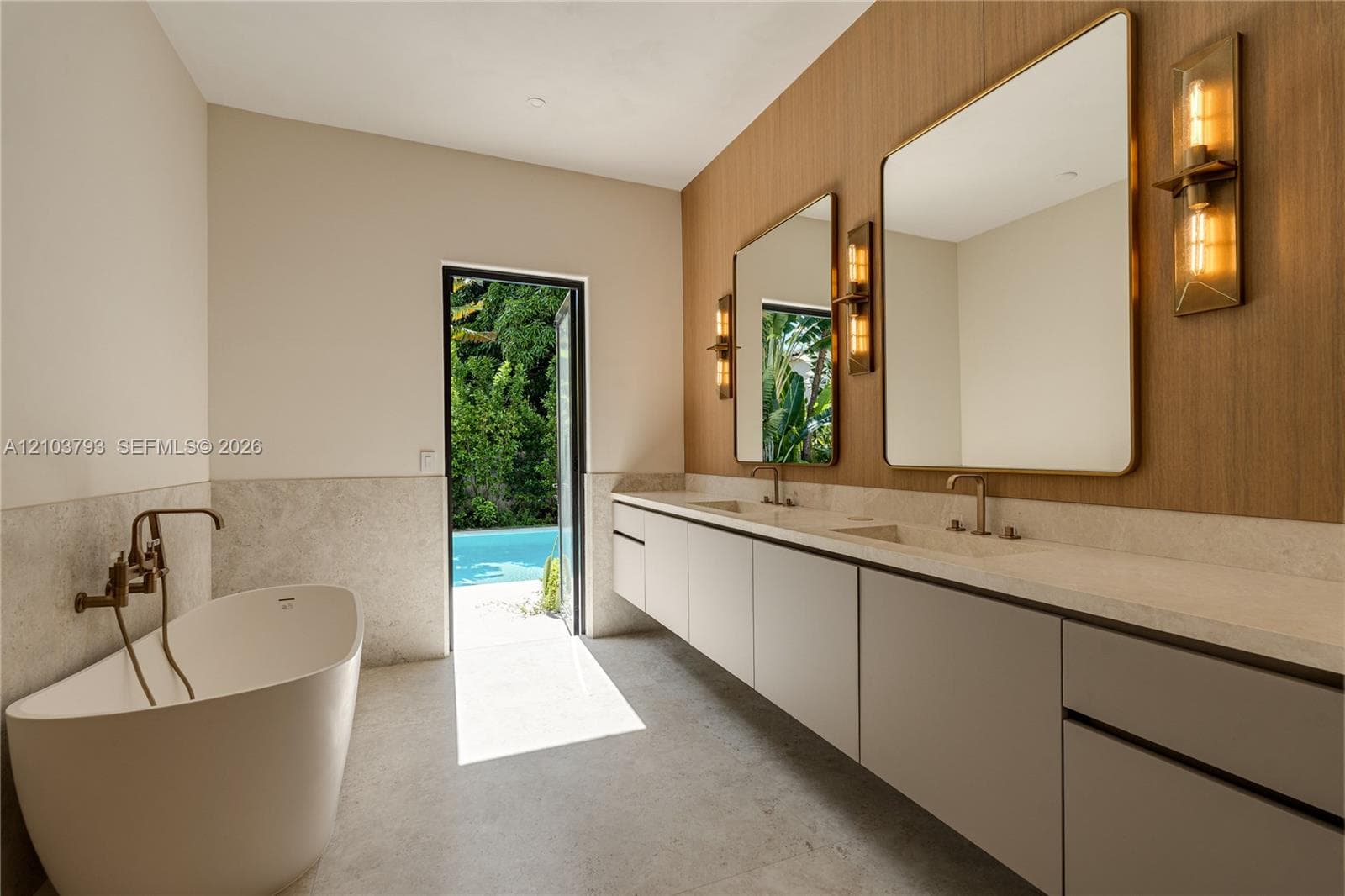 Floating double vanity with a light stone countertop, frameless mirrors with gold-tone sconces, freestanding soaking tub with floor-mounted faucet, and floor-to-ceiling stone wall tiling