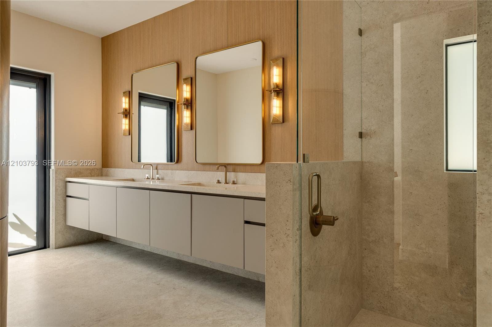 Freestanding soaking tub with wall-mounted metallic fixtures, floating dual-sink vanity with stone countertop, wood-paneled accent wall, rectangular framed mirrors, and metallic wall sconces