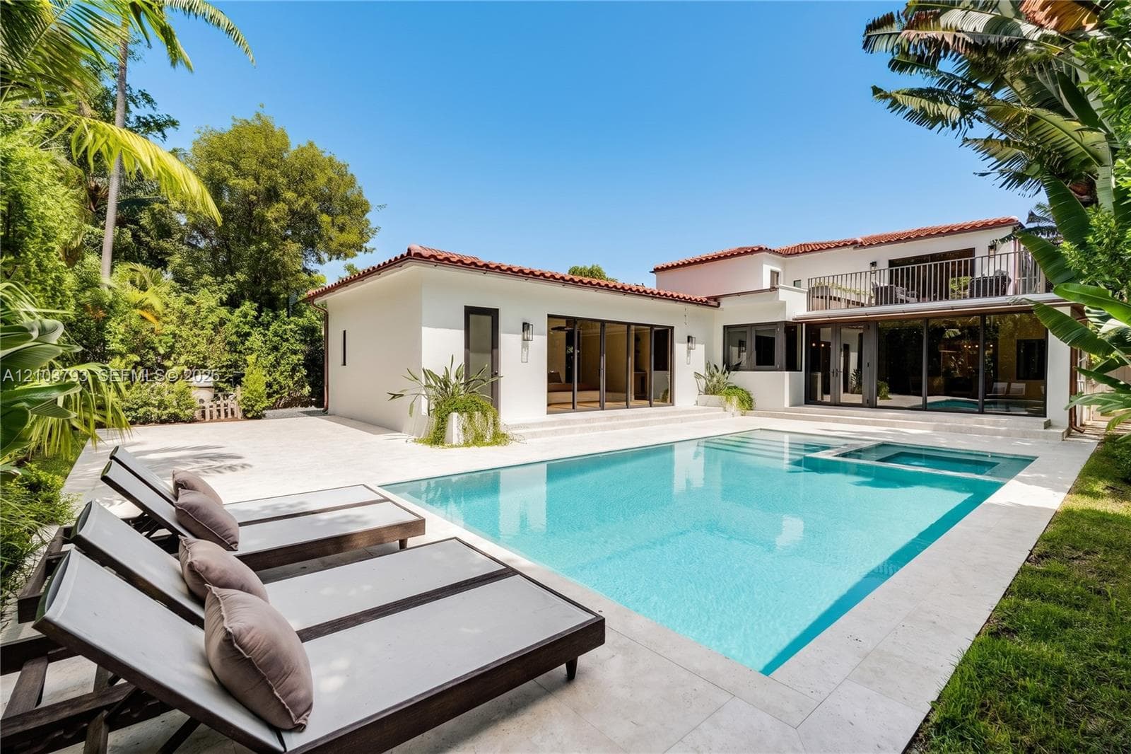 Exterior patio featuring a swimming pool with integrated spa, light-toned stone decking, multi-panel sliding glass doors, and a second-story metal railed balcony