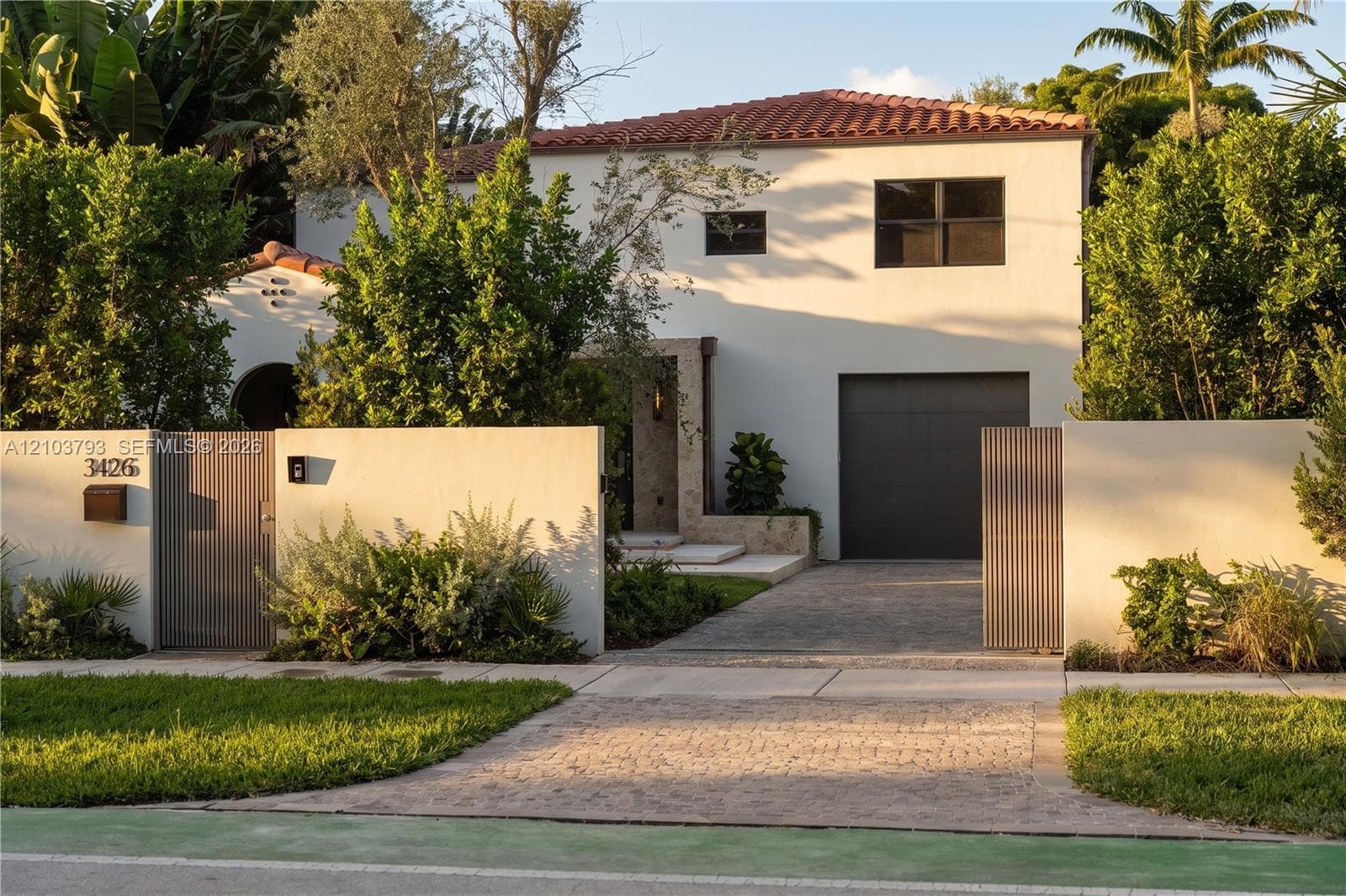 Two-story exterior featuring a smooth stucco finish, clay tile roof, stone-clad entrance, vertical slat privacy gates, and a paved stone driveway