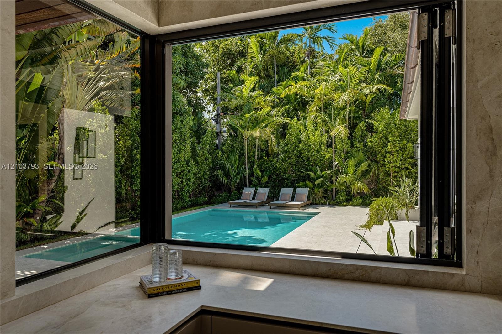 Corner floor-to-ceiling windows featuring black framing, stone countertop surfacing, and direct exposure to a swimming pool with stone decking and mature tropical landscaping