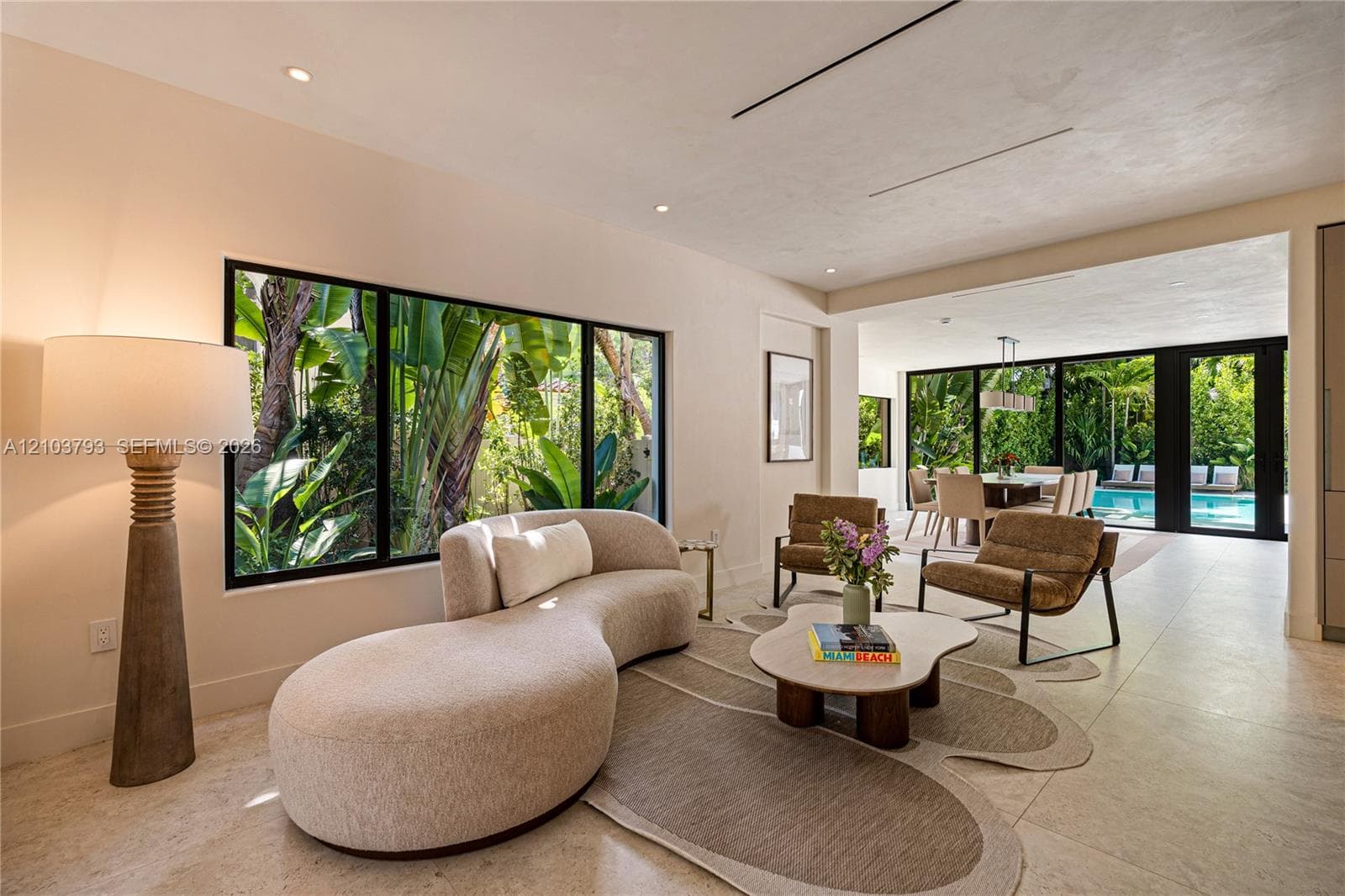 Interior space featuring large-format stone flooring, expansive black-framed windows, and recessed lighting, with retractable glass doors opening to an exterior swimming pool and lush landscaping